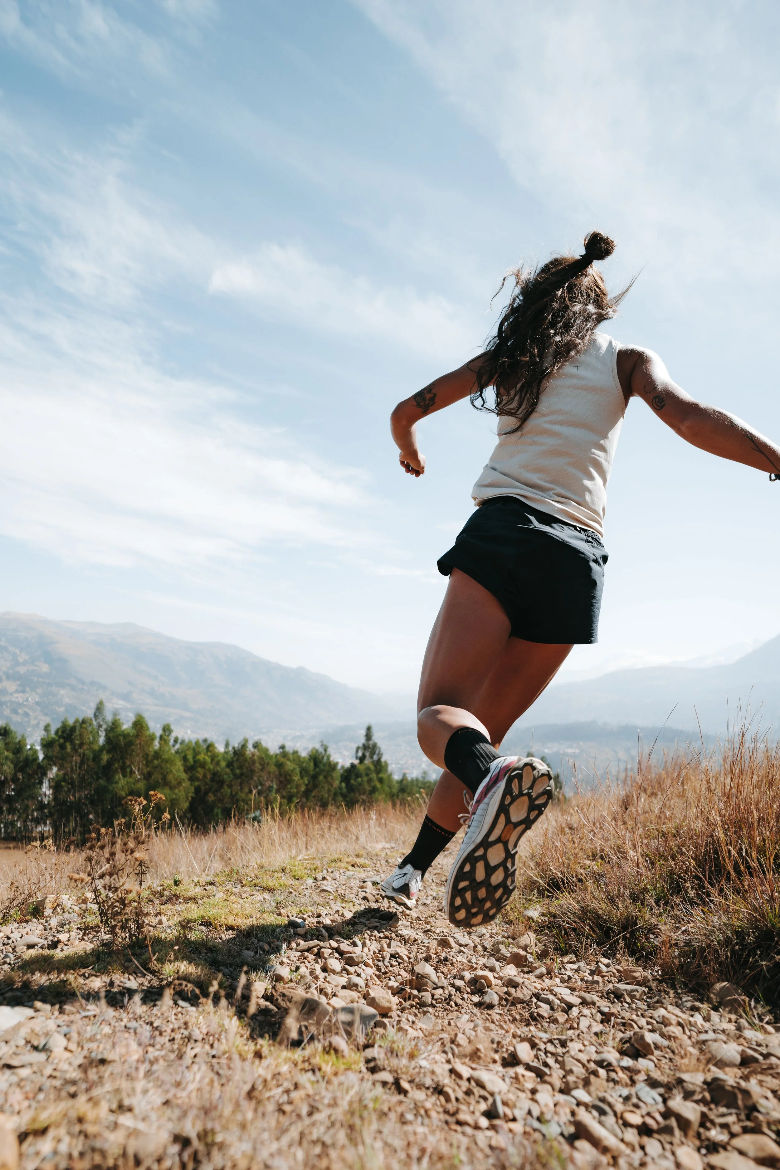 A woman running on a rocky trail in a mountainous outdoor setting, wearing athletic attire including a tank top, shorts, and trail running shoes.