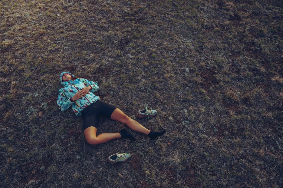A person lying on the ground in an outdoor setting, wearing a jacket and shorts, with sneakers off, appearing to rest or sleep.