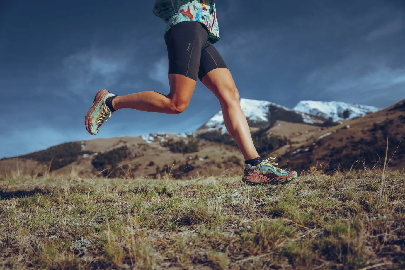 A person running outdoors on a grassy trail with mountains in the background, wearing athletic shoes, shorts, and a jacket.