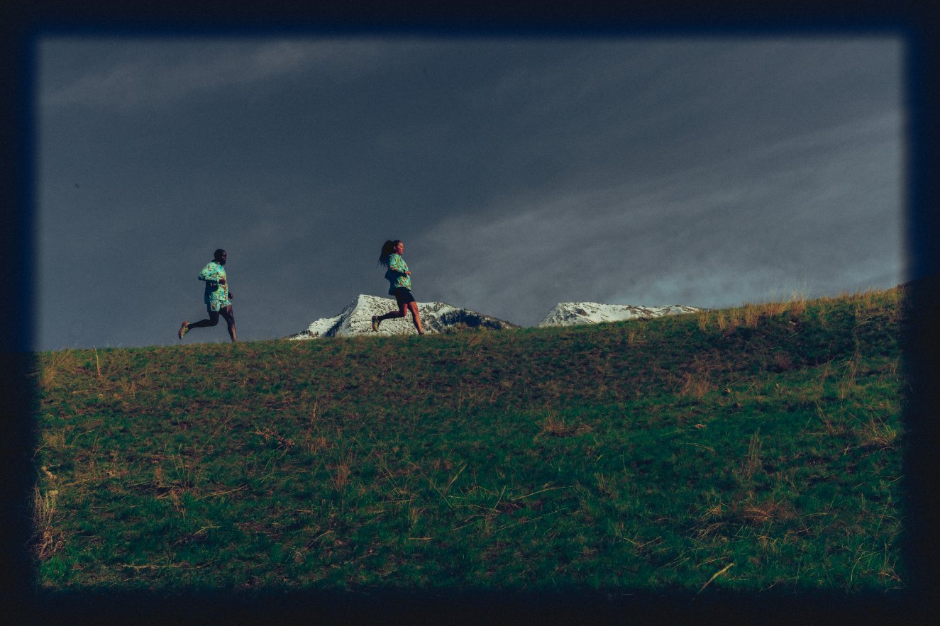 Two runners in sportswear running on a grassy hill with snow-capped mountains in the background under a cloudy sky.