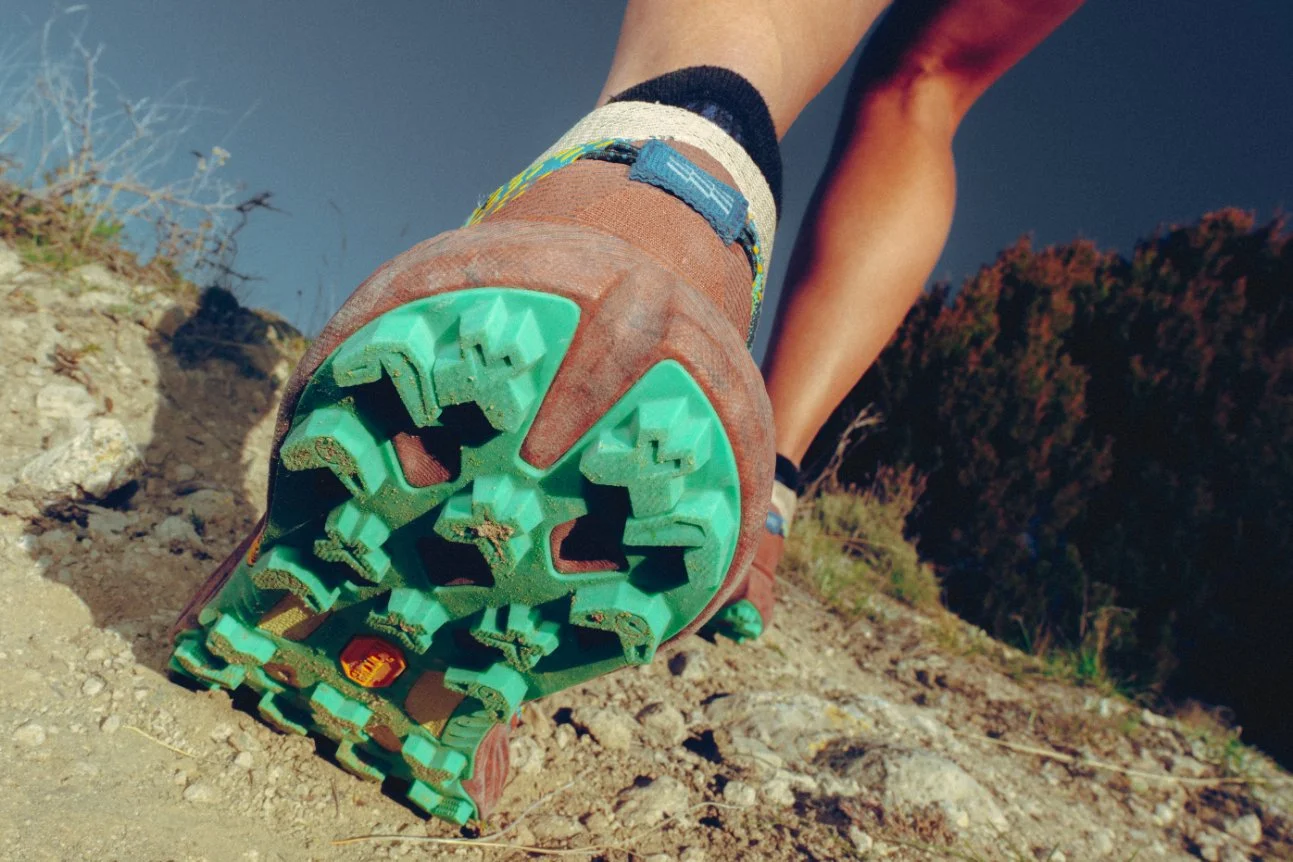 Close-up of a person's foot wearing a hiking shoe with a green and brown sole, walking on a dirt trail in a natural outdoor setting with shrubs and trees in the background.
