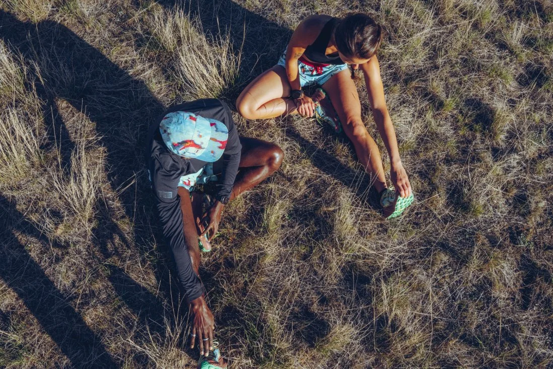 A woman and a young boy sitting on dry grass, tying their sneakers outdoors.