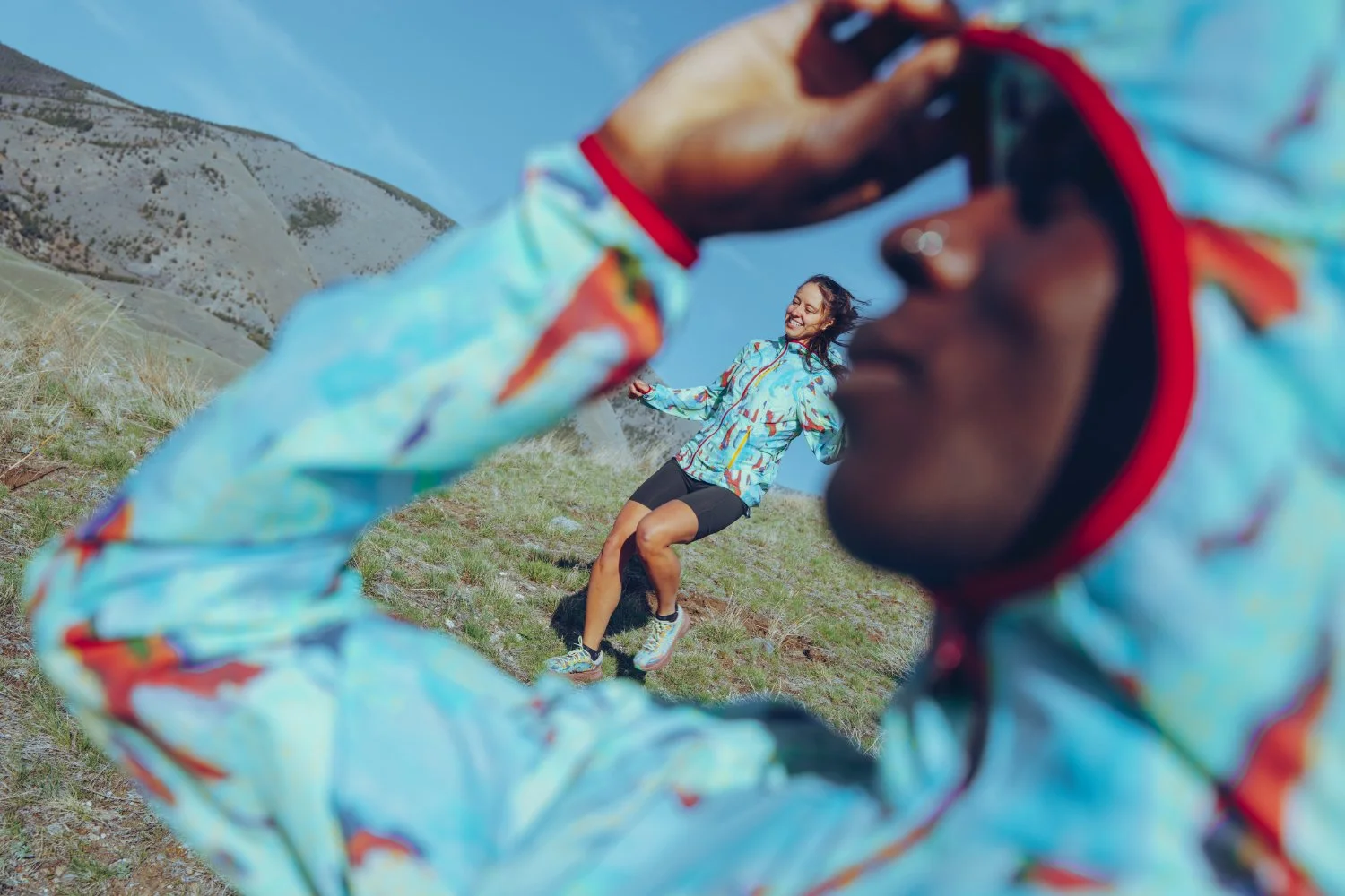 A woman in athletic clothing jumping joyfully on a hillside, as another person in colorful outdoor gear takes a photo with a camera.