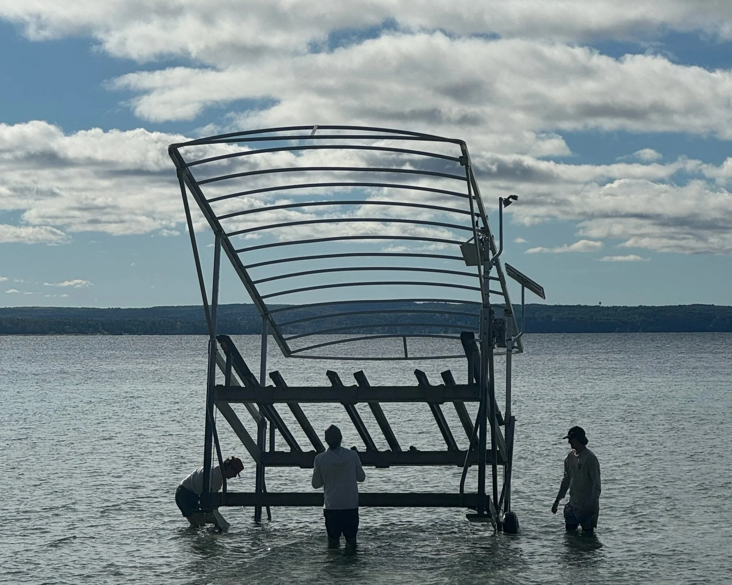 Three people standing in shallow water near a partially submerged large metal boat lift at the edge of a body of water, with a landscape of hills in the background under a partly cloudy sky.
