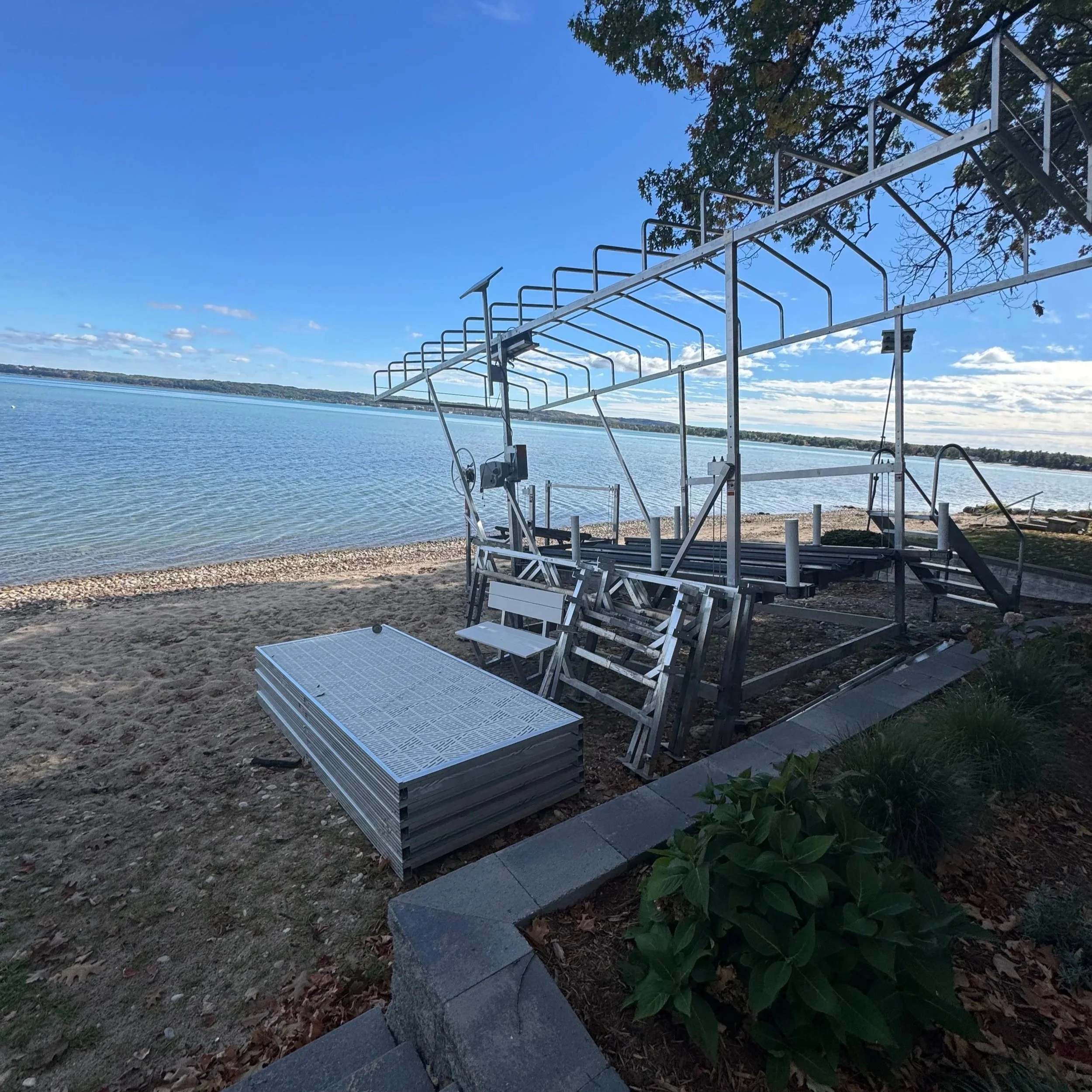 Dock and boat hoist on a beach near a body of water under a blue sky with scattered clouds, bordered by greenery and a concrete edge.