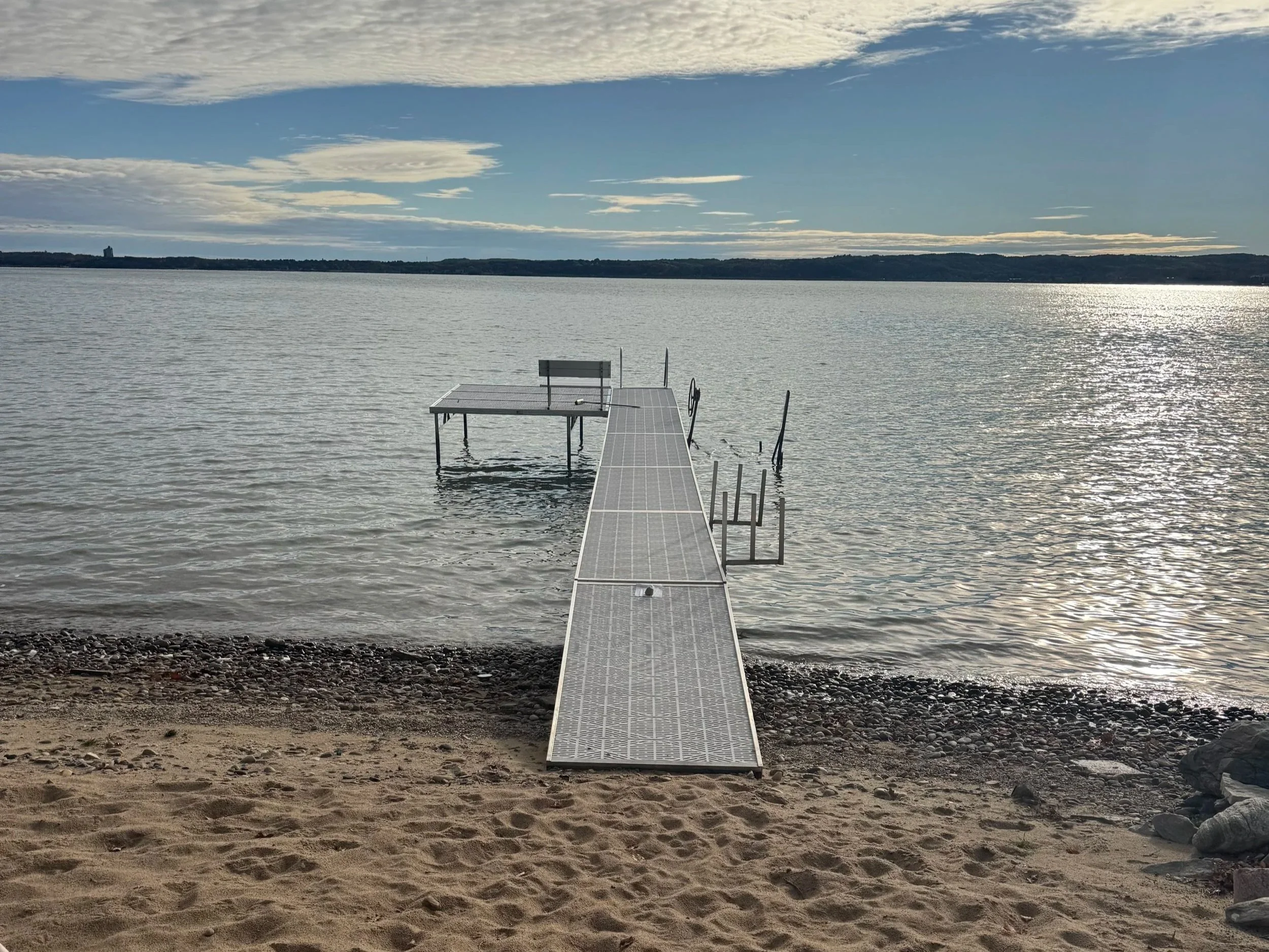 A professionally installed dock extending into a Michigan lake on a sandy beach, with cloudy sky and distant shoreline visible.