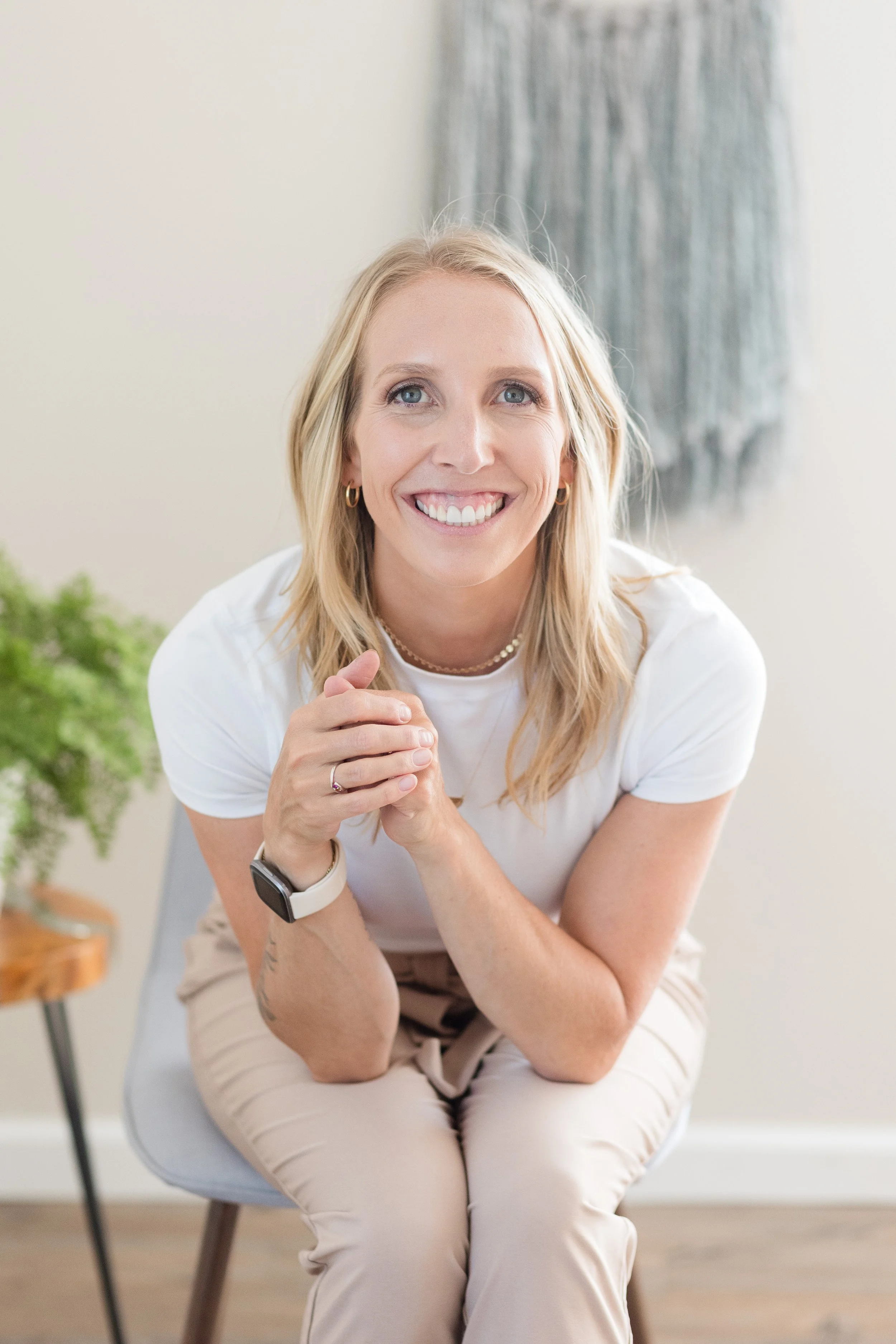 A woman sitting on a chair, smiling, with her hands clasped, wearing a white t-shirt, beige pants, a watch, and jewelry, in a bright, minimal indoor setting.