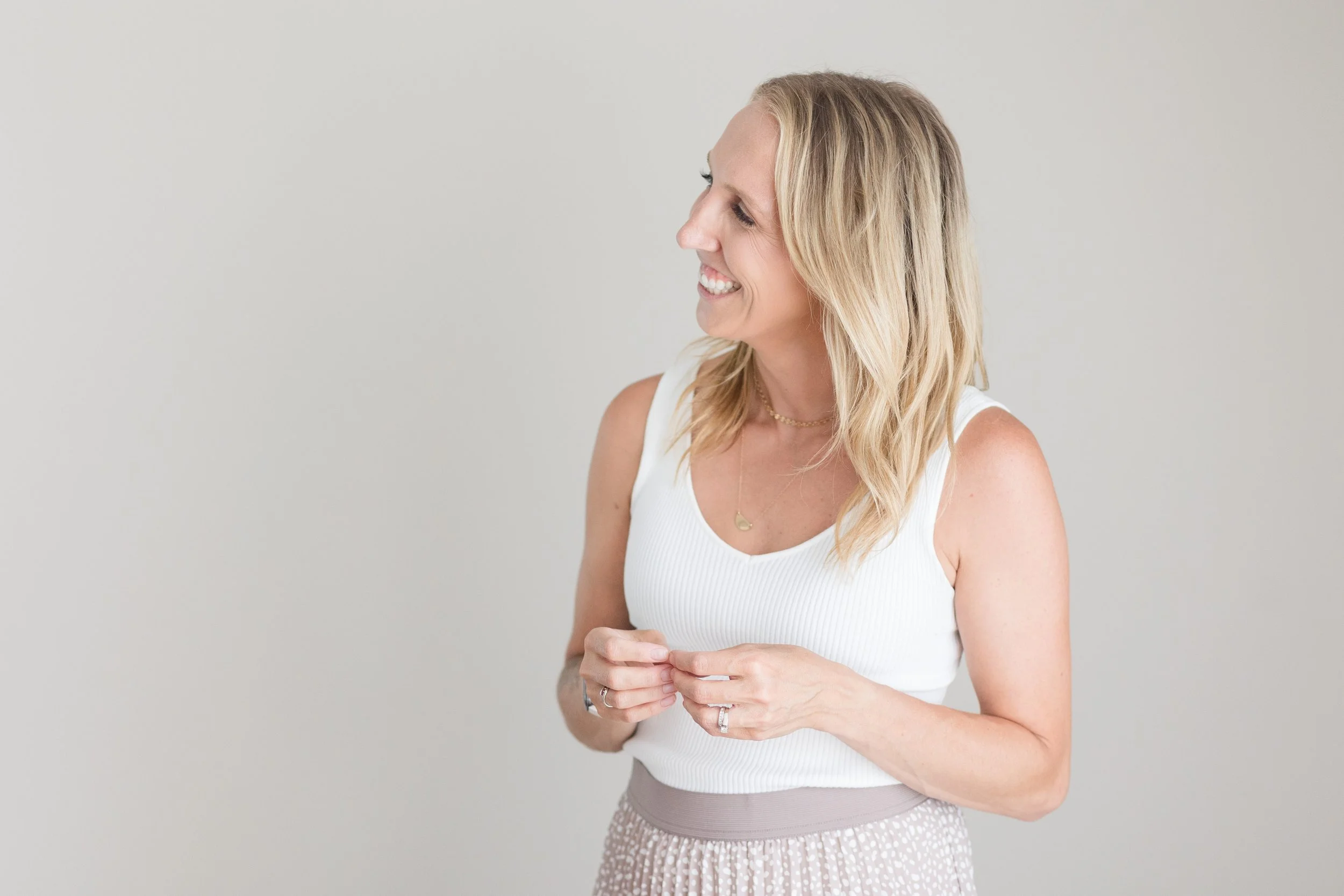 A woman with blonde hair smiling and looking to her left, wearing a white sleeveless top and a patterned skirt, holding her hands in front of her