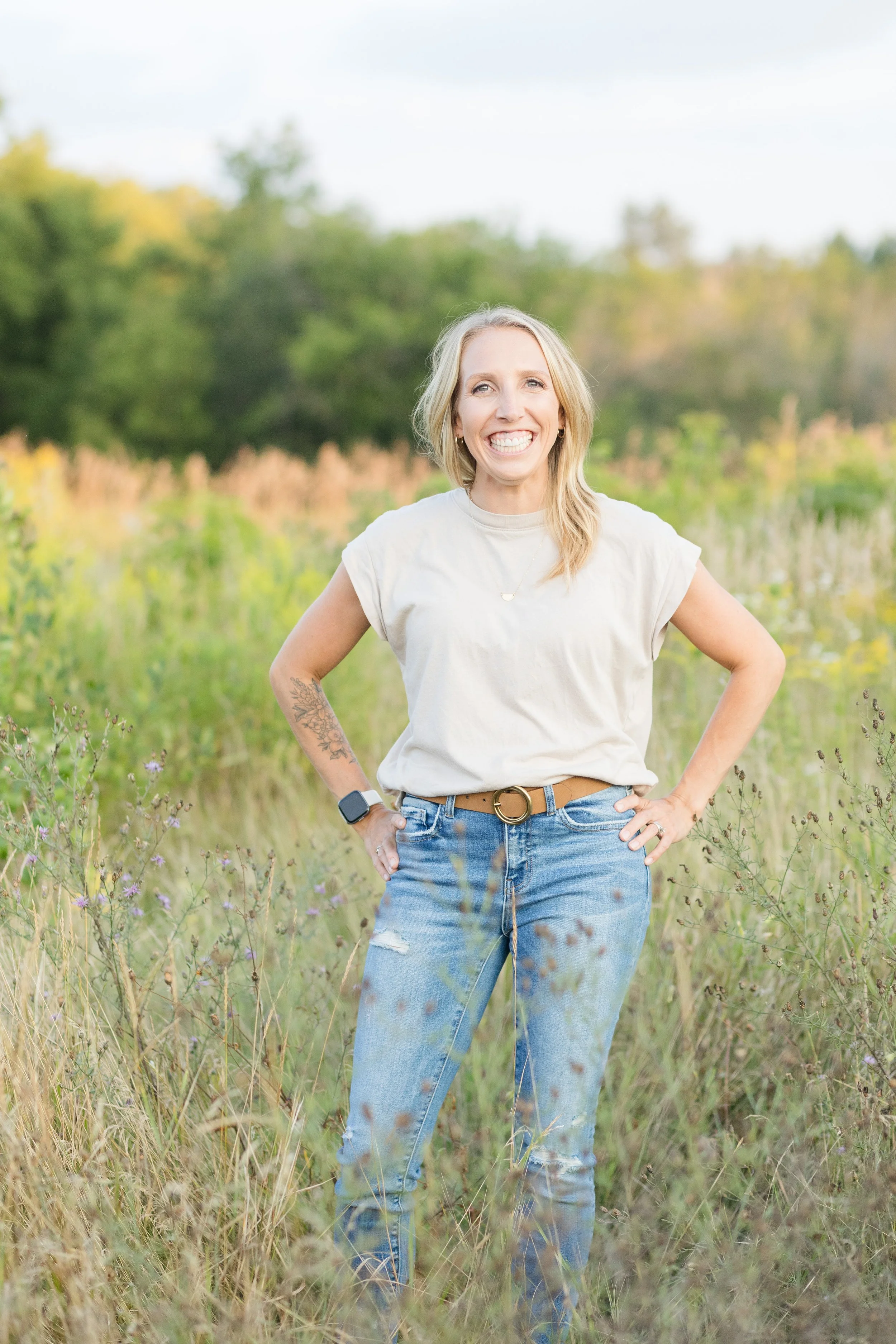 A woman standing outdoors in a grassy field with trees in the background, smiling at the camera.