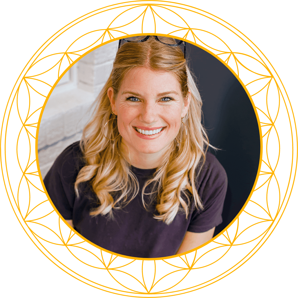 A woman with long blonde hair, blue eyes, and glasses on her head, smiling in a close-up photo with a black background and gold geometric circular designs.