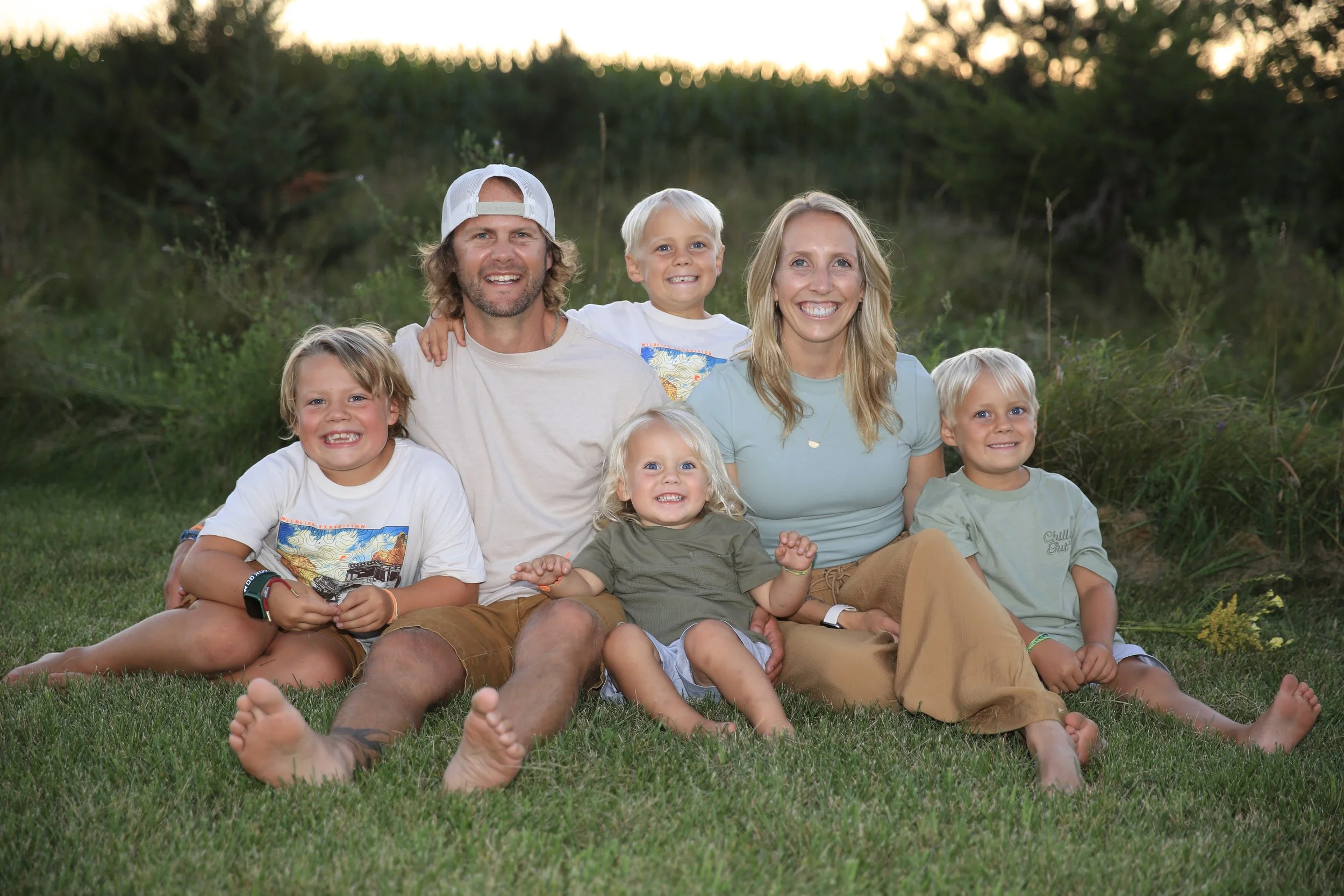 Family of six sitting on the grass outdoors, smiling at sunset. Three children, two adults, natural background of trees and bushes.
