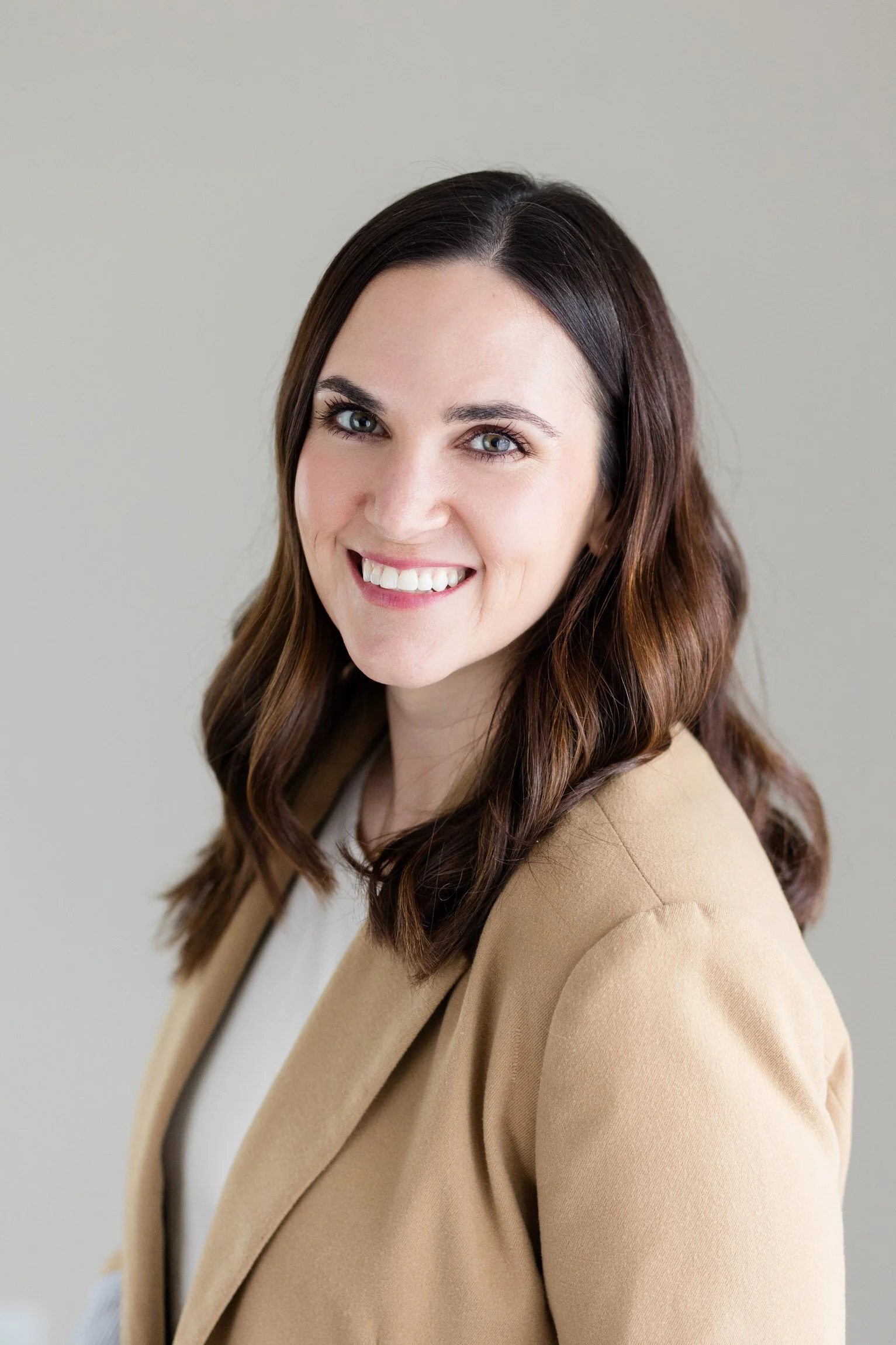 A woman with brown hair and blue eyes smiling while looking at the camera, wearing a beige blazer and a white top, against a plain light background.