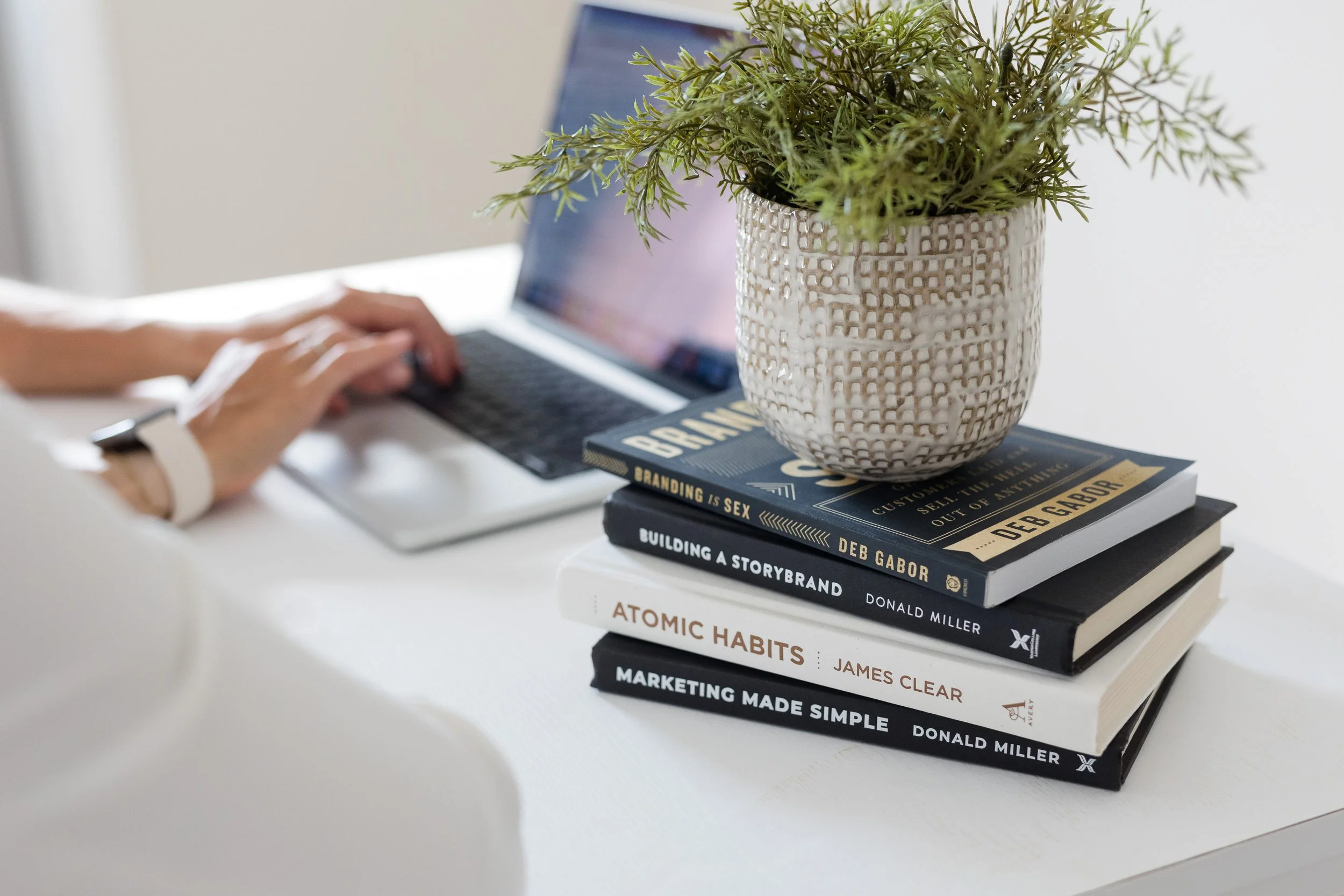 A stack of five books labeled 'Branding is Sex,' 'Building a StoryBrand,' 'Atomic Habits,' and 'Marketing Made Simple,' topped with a potted green plant, on a white desk. A person's hand is typing on a laptop in the background.