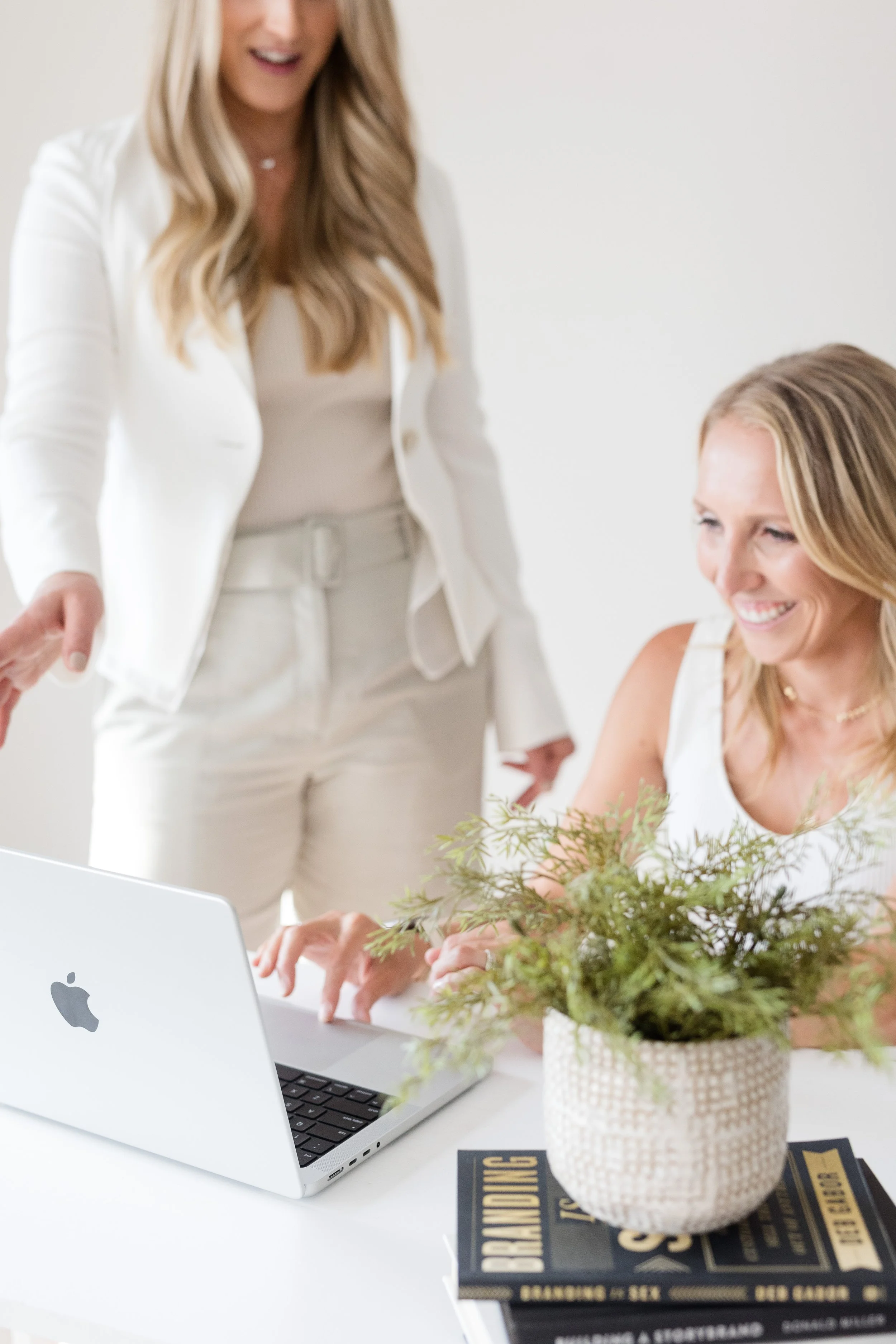 Two women working at a laptop in a bright, modern office. One woman is sitting and smiling while the other stands near her. There is a potted plant and books on the table.