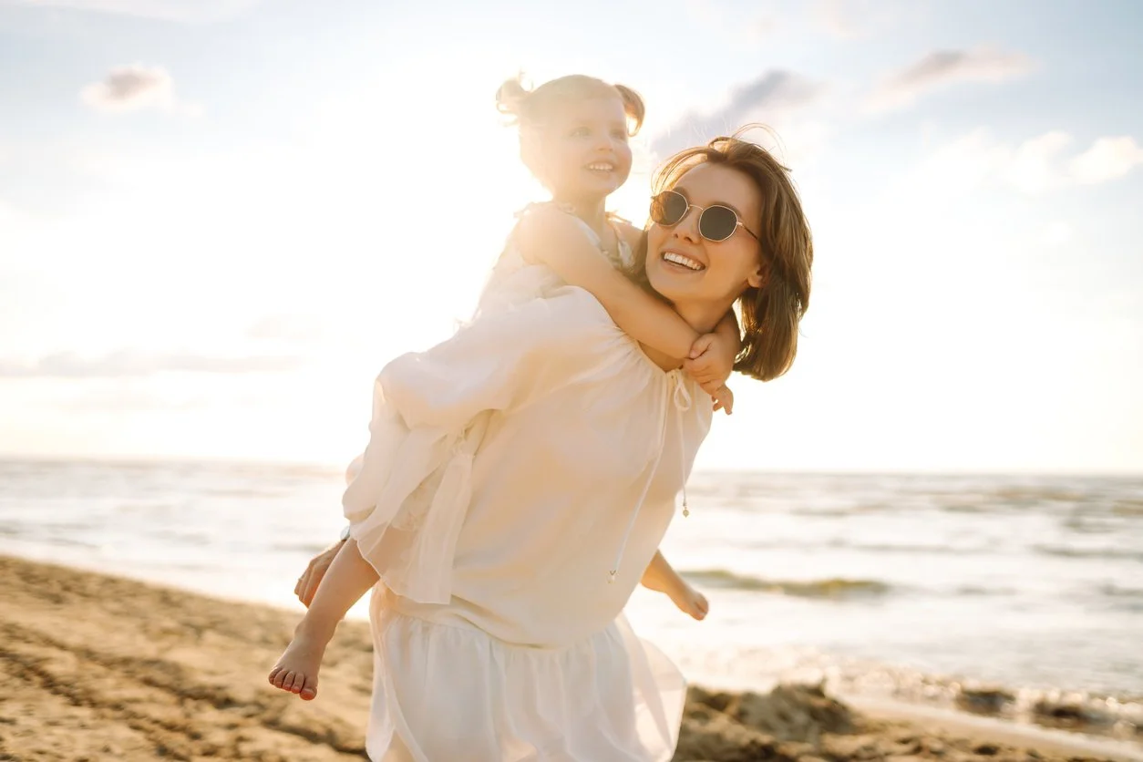 A woman wearing sunglasses carries a young girl on her back at the beach while working on speech and language skills in San Clemente, CA.