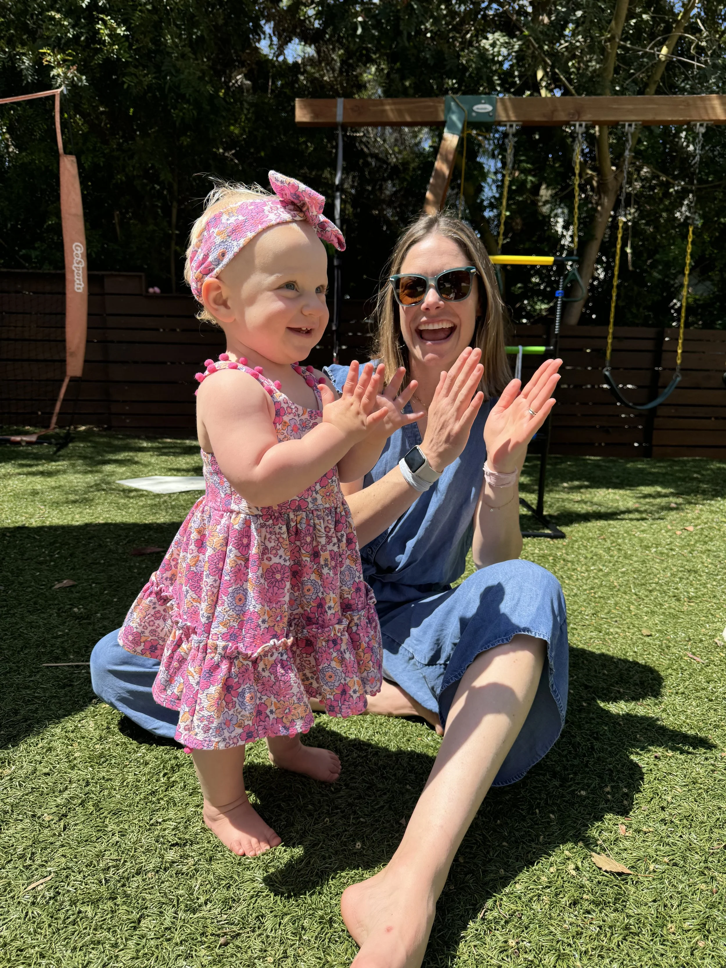 Jamie Montgomery, owner and pediatric speech therapist at Waves of Speech in San Clemente, CA, sitting cross-legged on the grass, smiling and clapping with a young girl in a floral dress during a speech therapy session.
