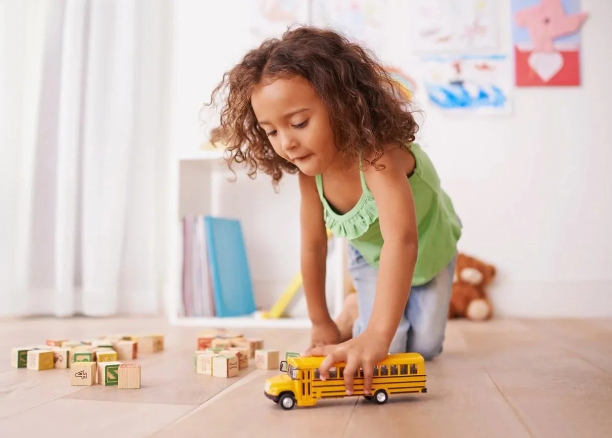 A young girl with curly hair, playing on the floor with toy wooden blocks and a yellow school bus during speech therapy for kids in San Clemente, CA.