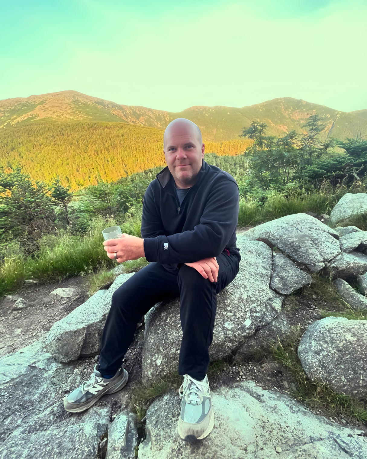 A man sitting on rocks outdoors with mountains and green trees in the background, holding a glass, during daytime.