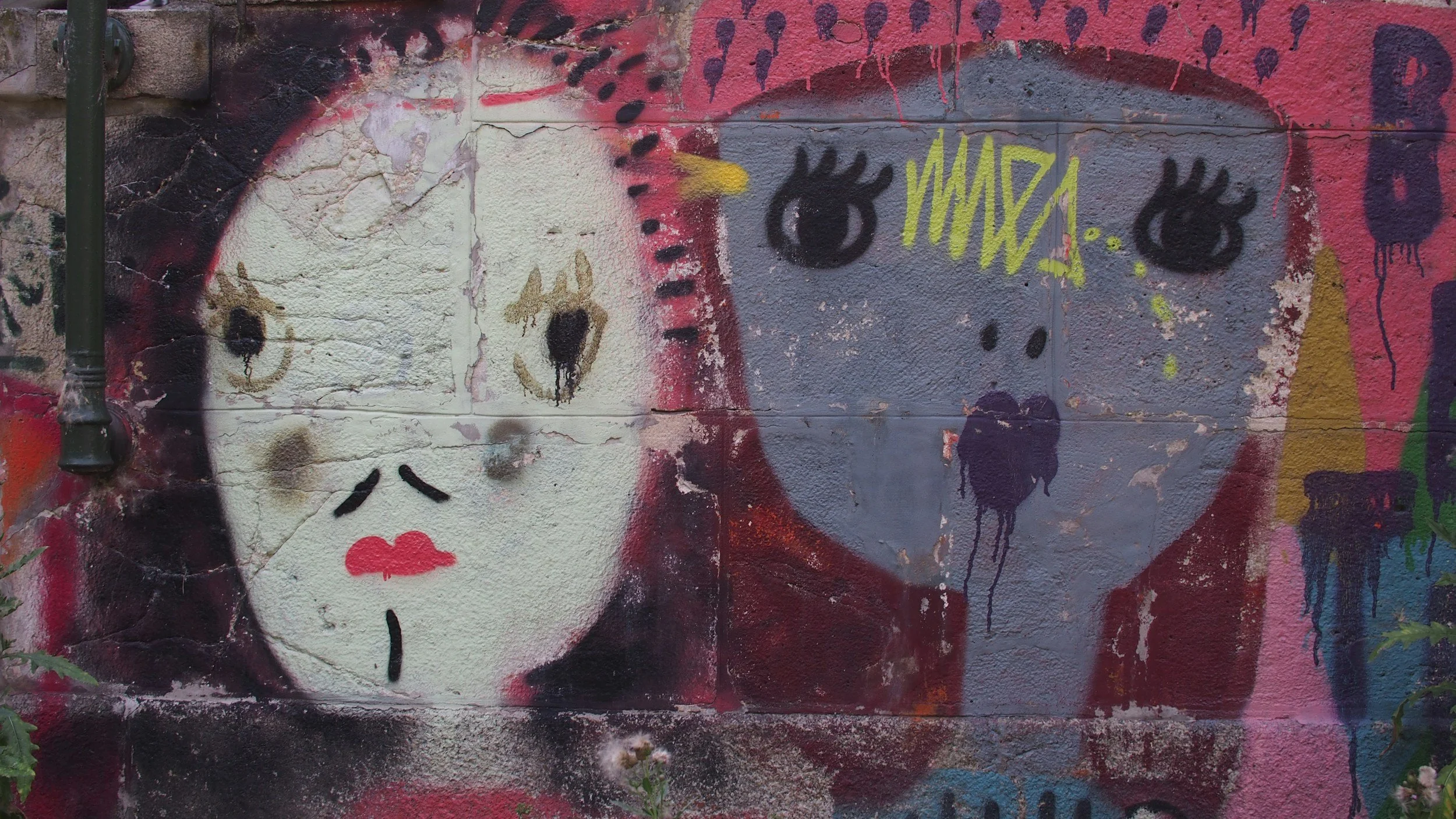 Graffiti of a sad woman's face with black hair and pink lips, next to a gray face with closed eyes and yellow graffiti writing above it, on a textured wall.