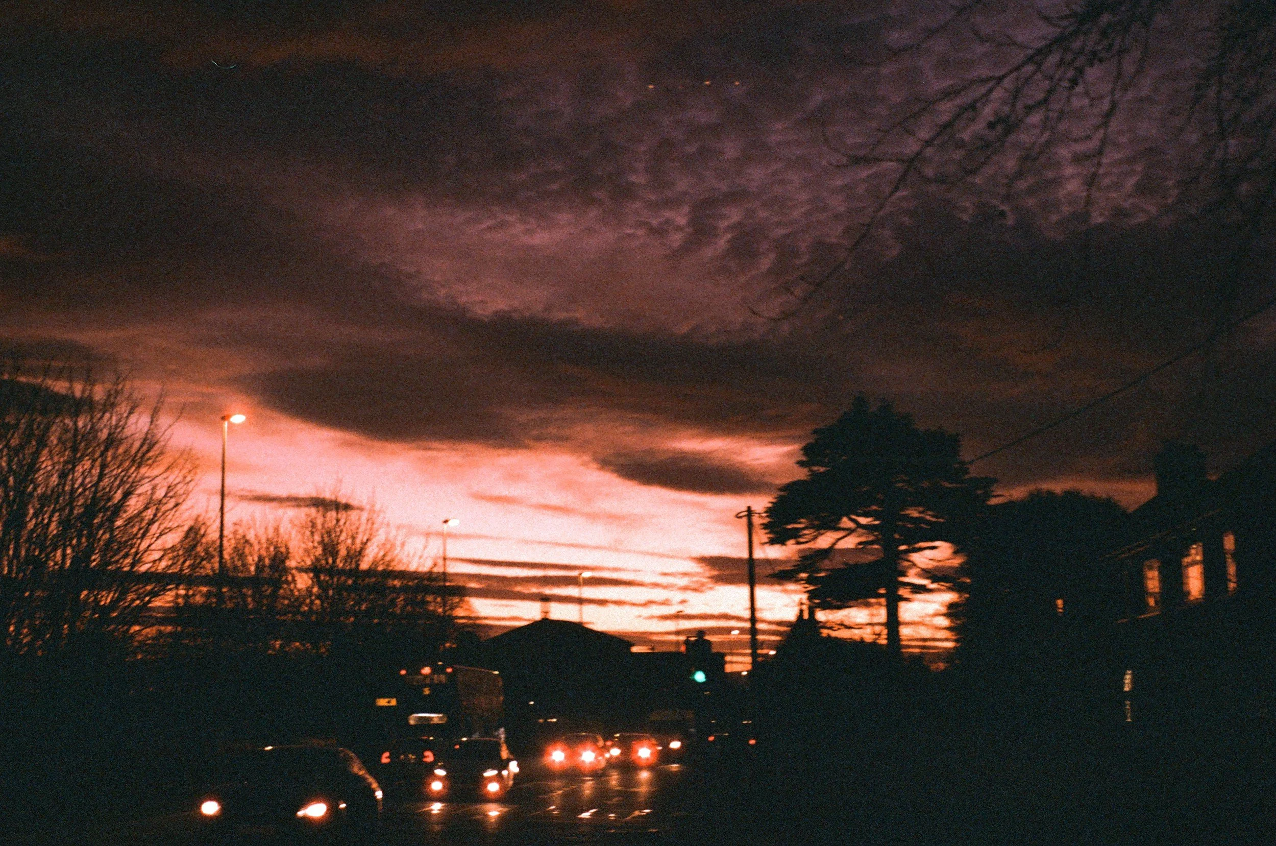 Darkening sky with clouds during sunset, silhouetted trees and buildings, streetlights, and cars with headlights on
