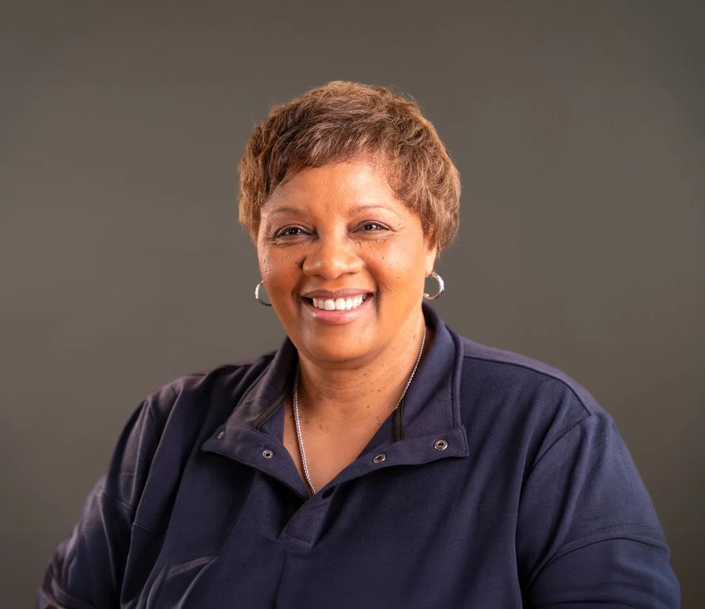 A woman with short brown hair, smiling, wearing a dark navy jacket and hoop earrings, against a plain gray background.