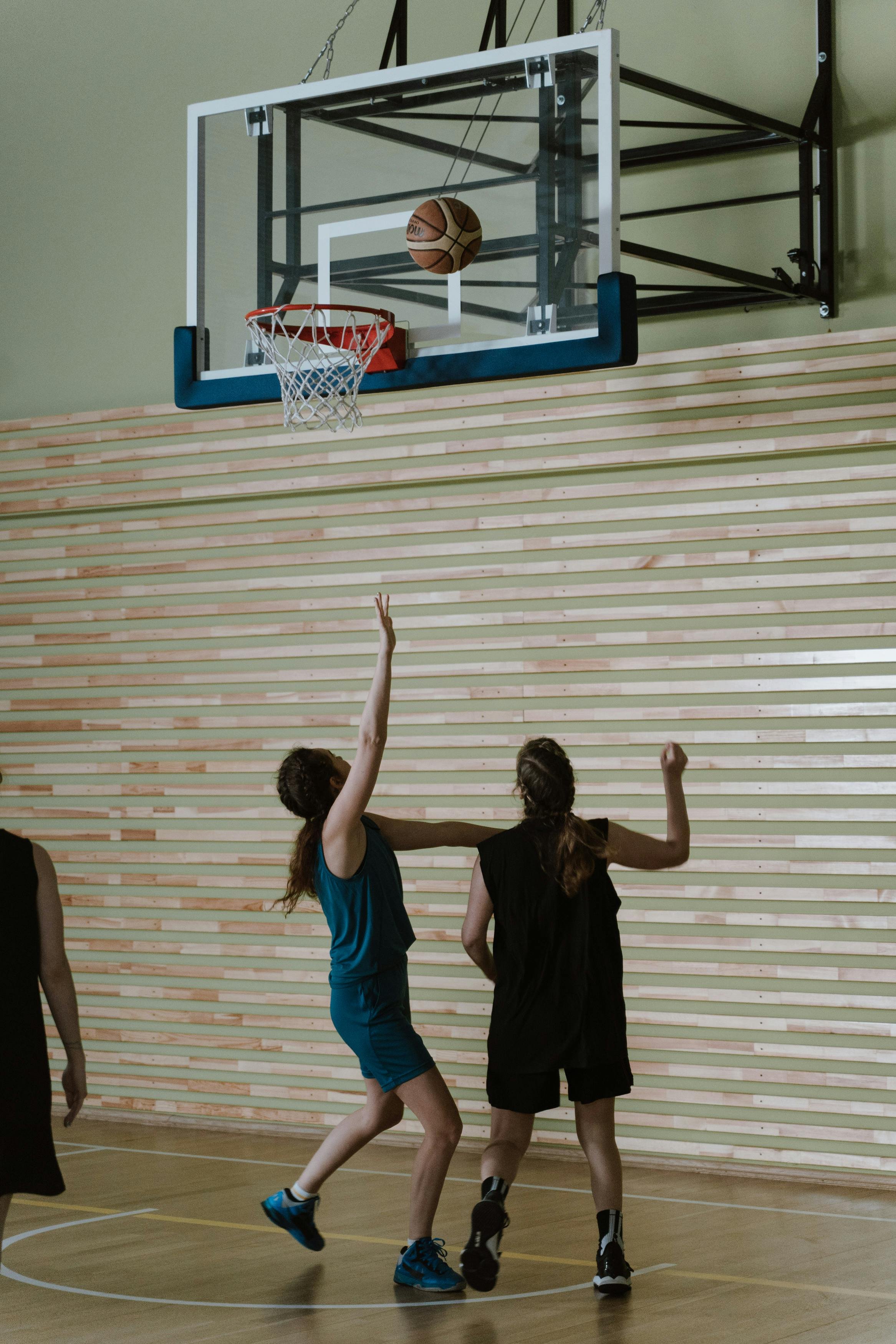 Girls playing basketball in an indoor gym, reaching up towards the basketball hoop as the basketball is about to go through the hoop.