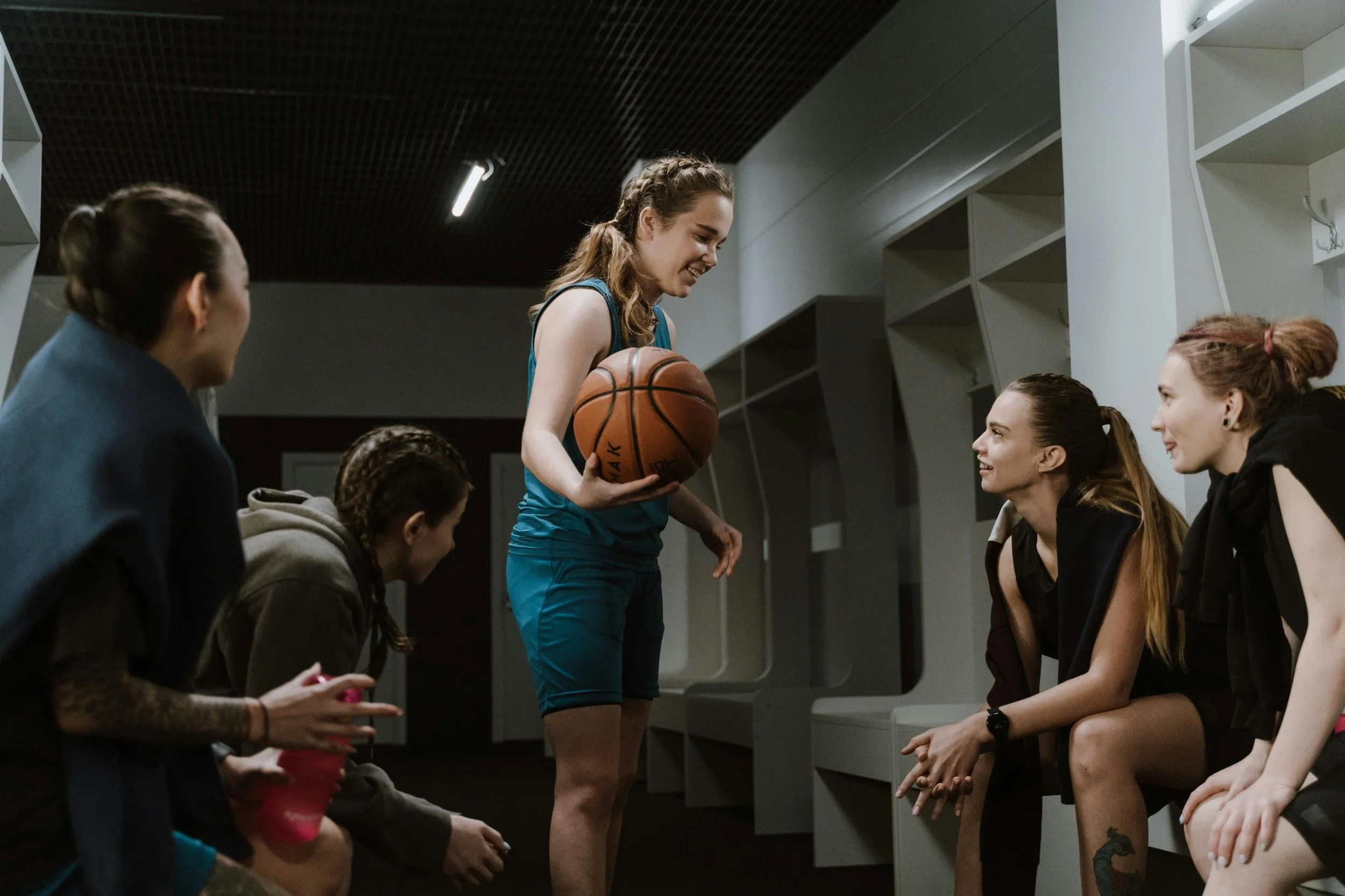 A female basketball player in teal uniform holding a basketball while talking to a group of women sitting on benches in a locker room, with some women smiling and listening.