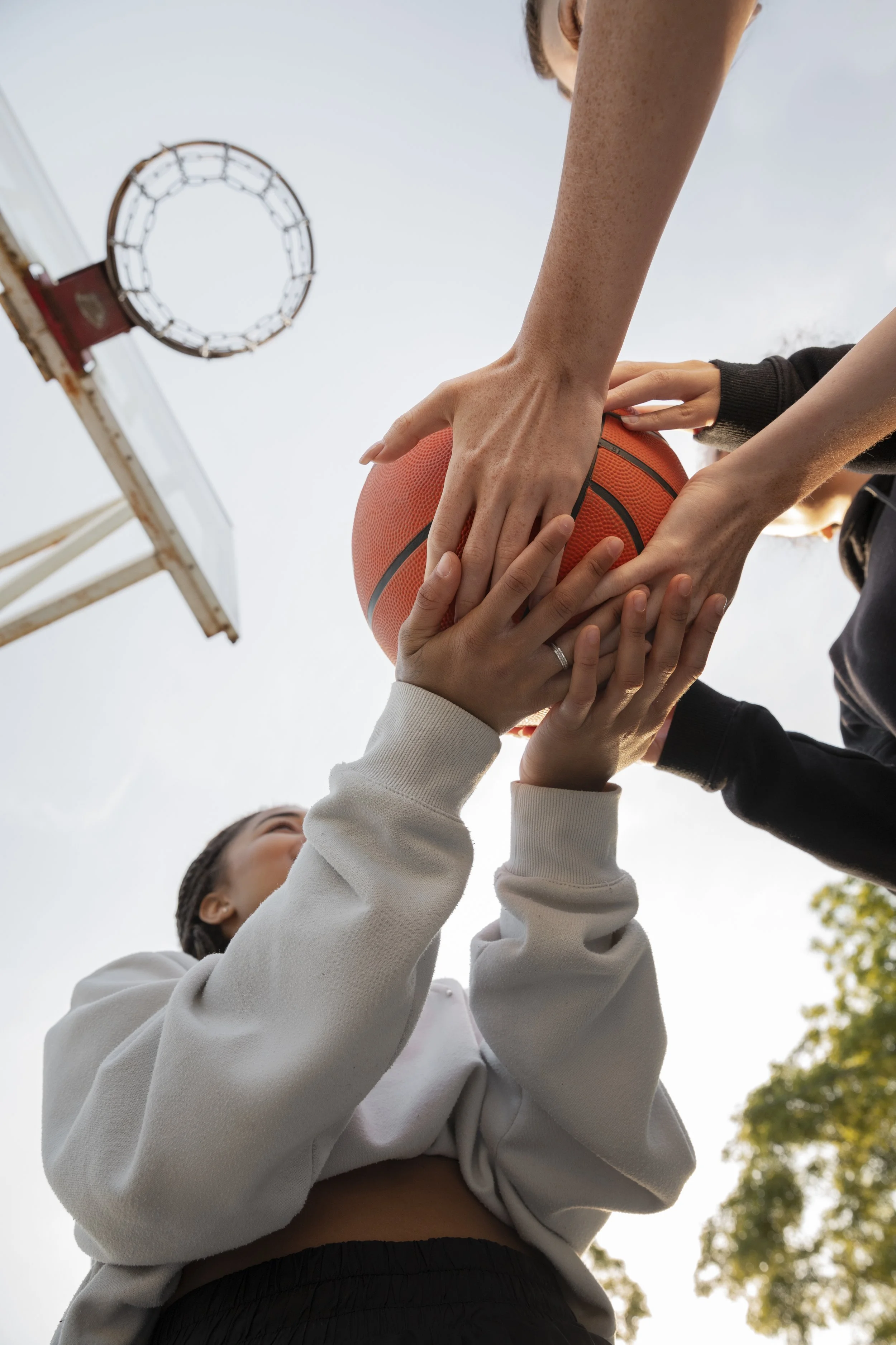 Close-up of people playing basketball outdoors, with hands on the ball and a basketball hoop visible in the background on a clear day.