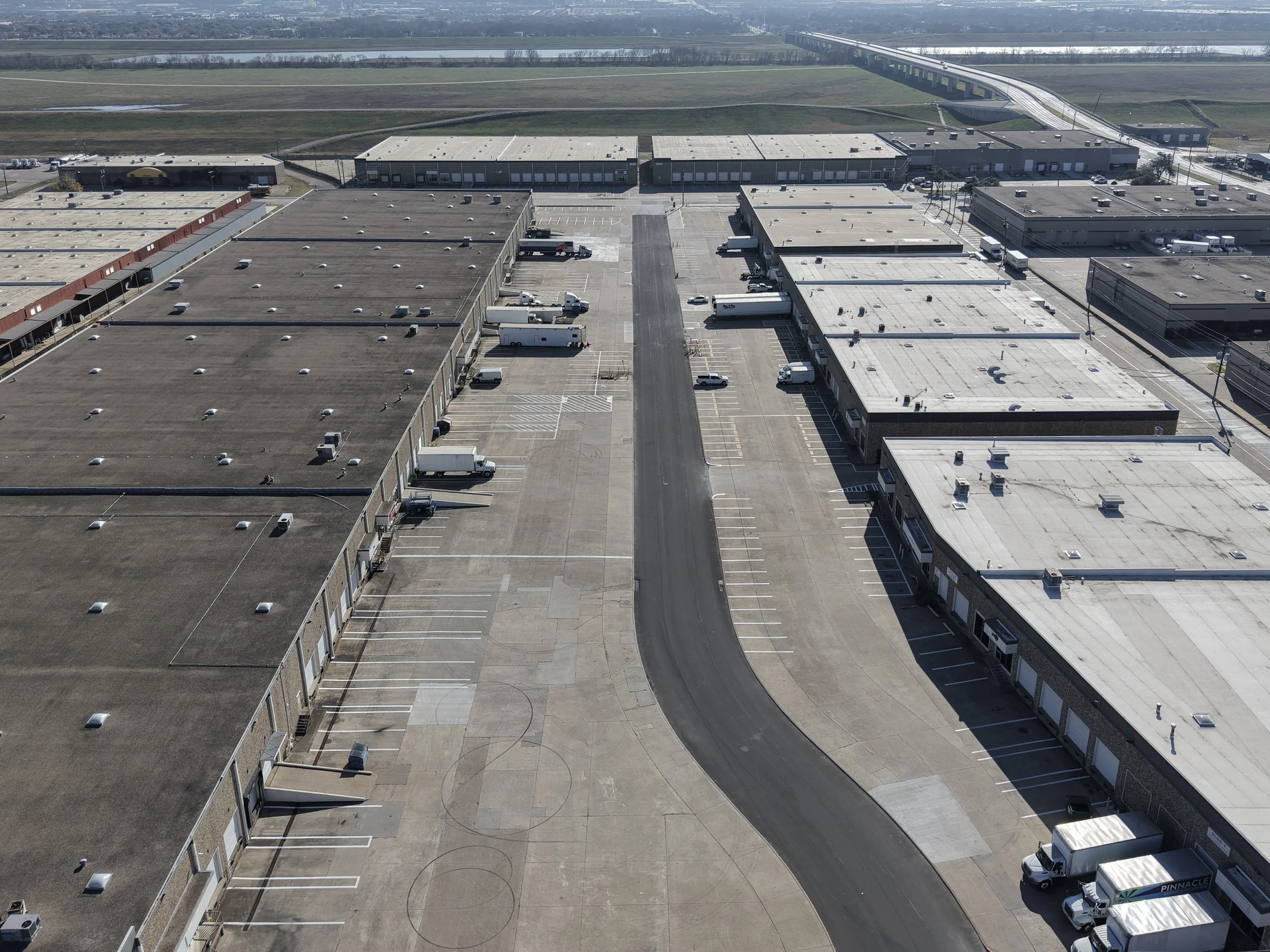 An aerial view of a large warehouse complex with a parking lot, loading docks, and empty parking spaces. A curved road runs through the warehouse area. In the background, there are fields and a highway.