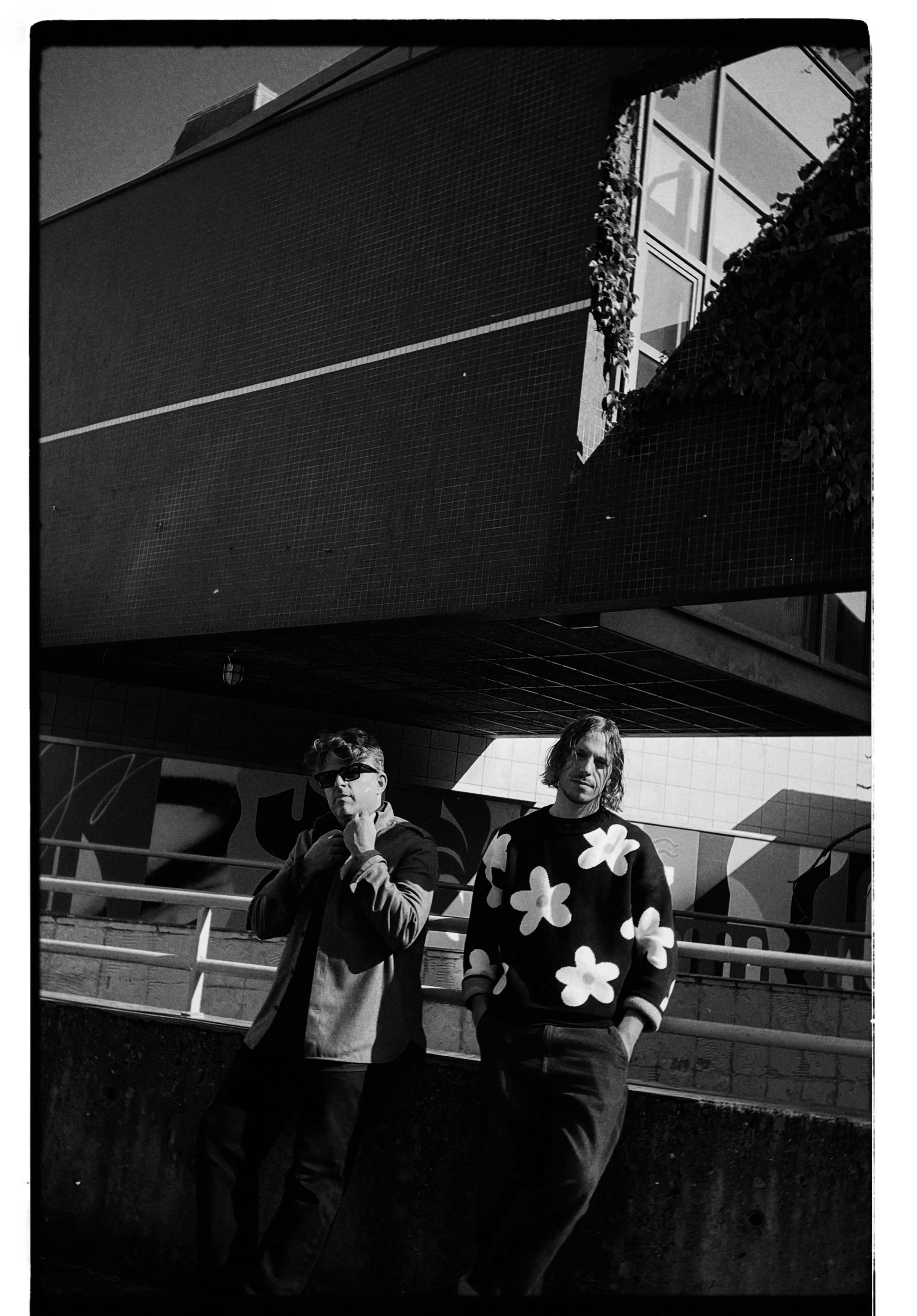 Two men standing outdoors in front of a modern building with large windows, black and white photo. One man is wearing sunglasses and a two-tone jacket, the other has shoulder-length hair and a sweater with large flower patterns.