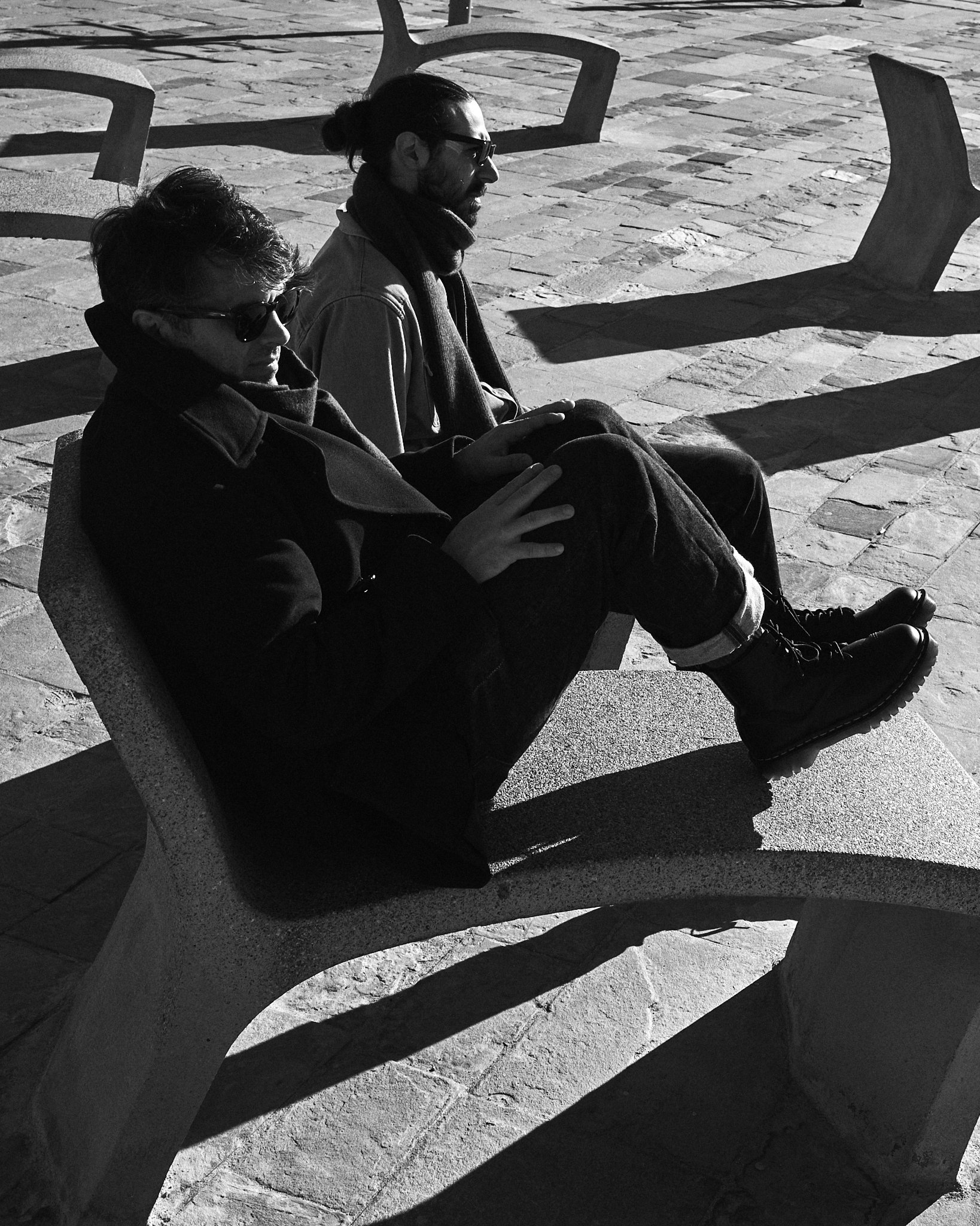 Two people sitting on a concrete park bench, wearing dark clothing and sunglasses, in bright sunlight, with shadows cast on the ground.