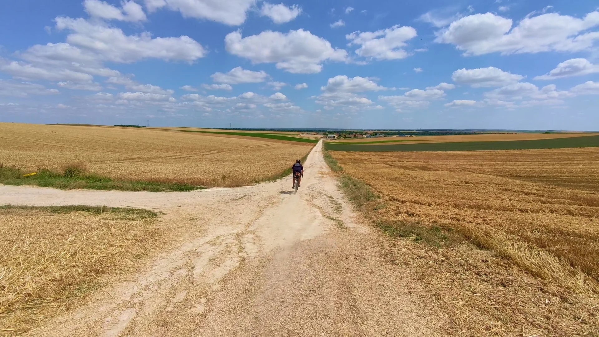 A person cycling on a dirt road through open fields under a blue sky with scattered clouds.