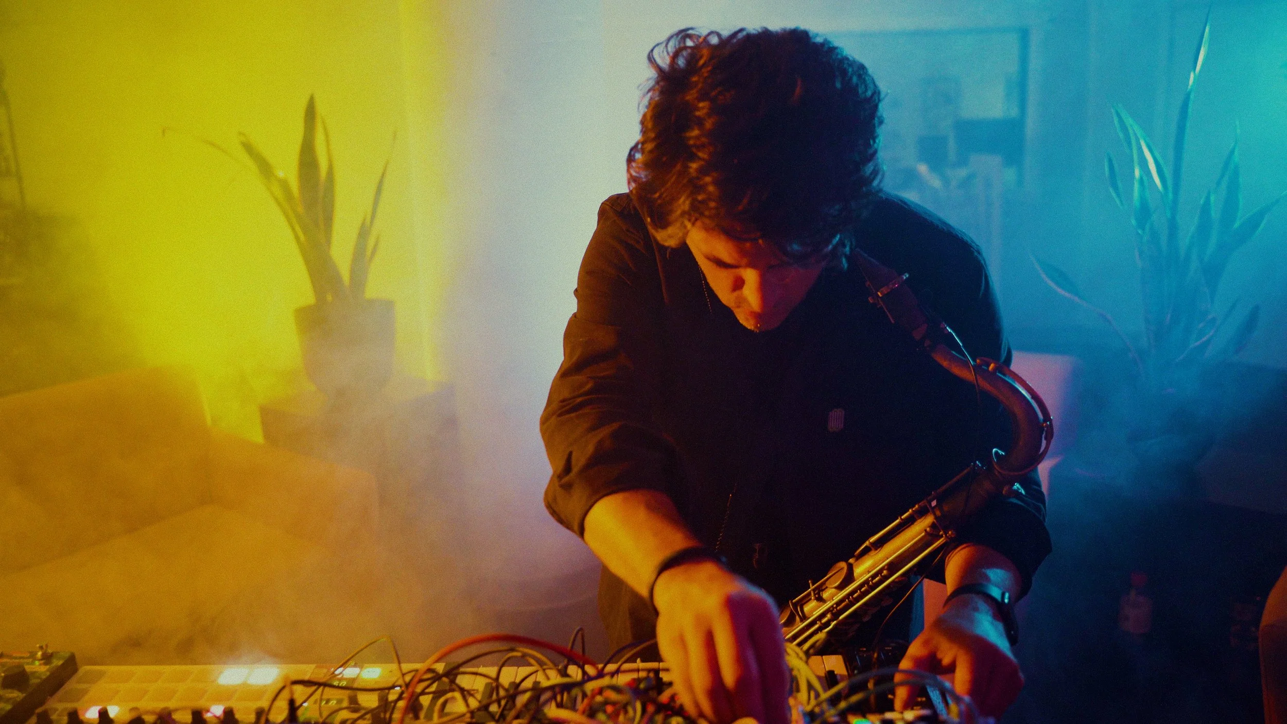 Person with curly hair playing a saxophone at a music setup with colorful lighting and fog effects.