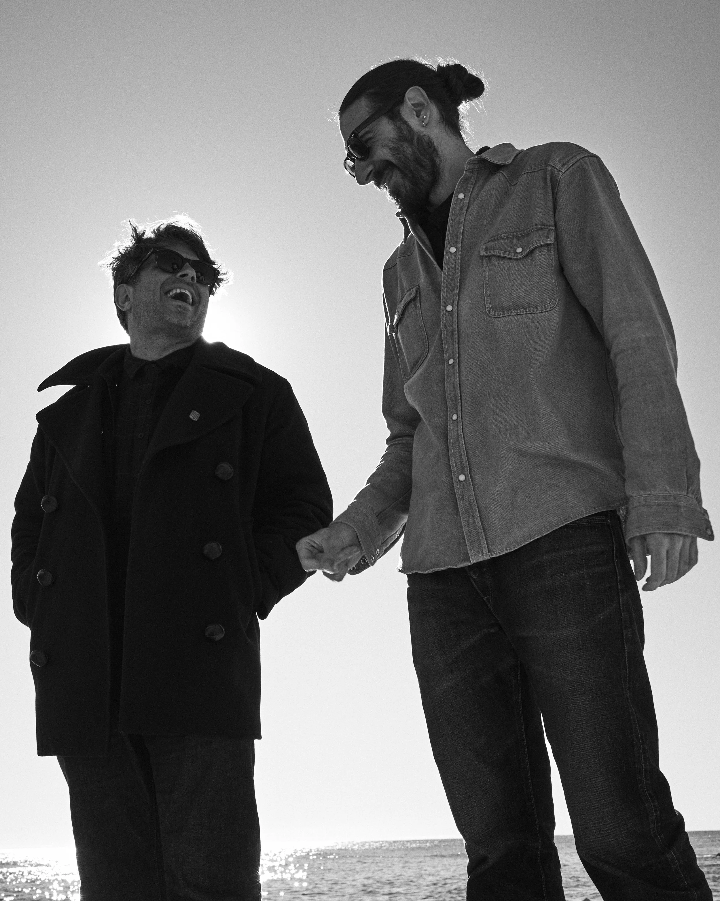 Two men with beards and long hair, wearing sunglasses, are smiling and holding hands outdoors near the water in black and white.
