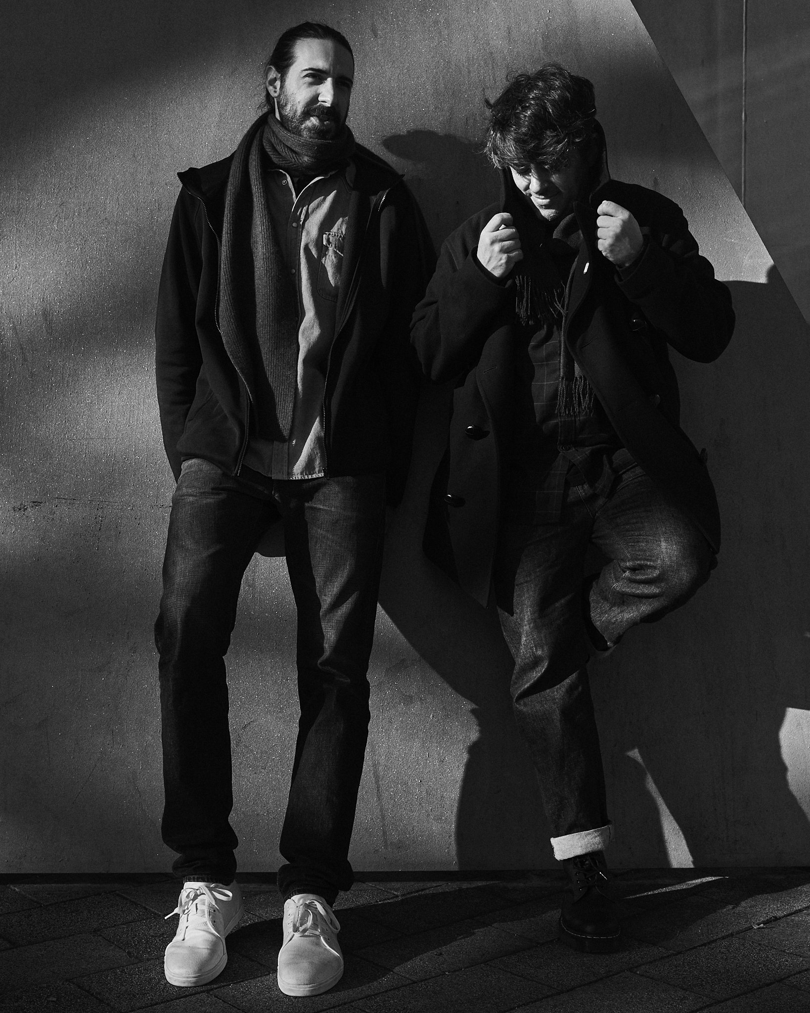 Two men standing against a wall in black and white, one with a beard and the other with a full head of hair, both dressed in casual winter clothing, with one man leaning against the wall.