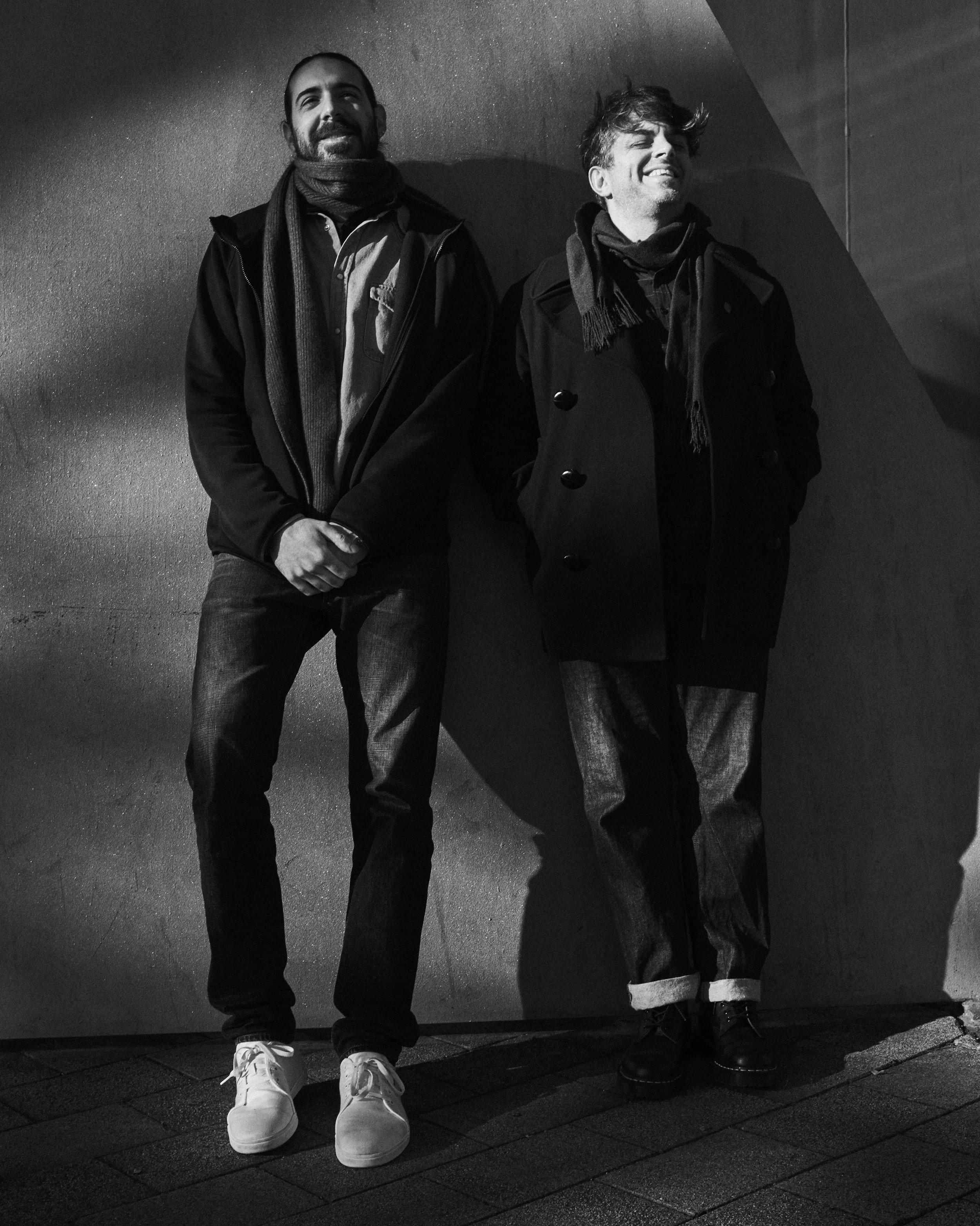 Two men standing against a textured wall, smiling and laughing, dressed warmly in jackets and scarves, with shadows cast on the wall.