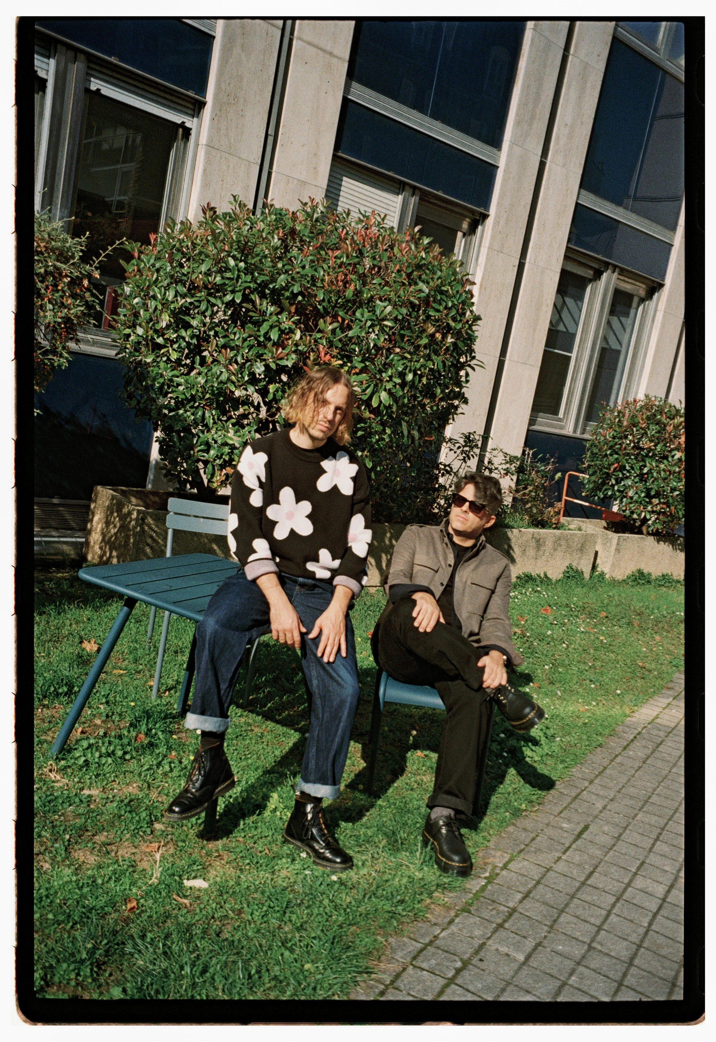 Two people sitting outside on chairs on grass next to a building with large windows and bushes. One person is wearing a black sweater with large white flowers, jeans, and black boots, while the other is wearing sunglasses, a gray jacket, black pants,