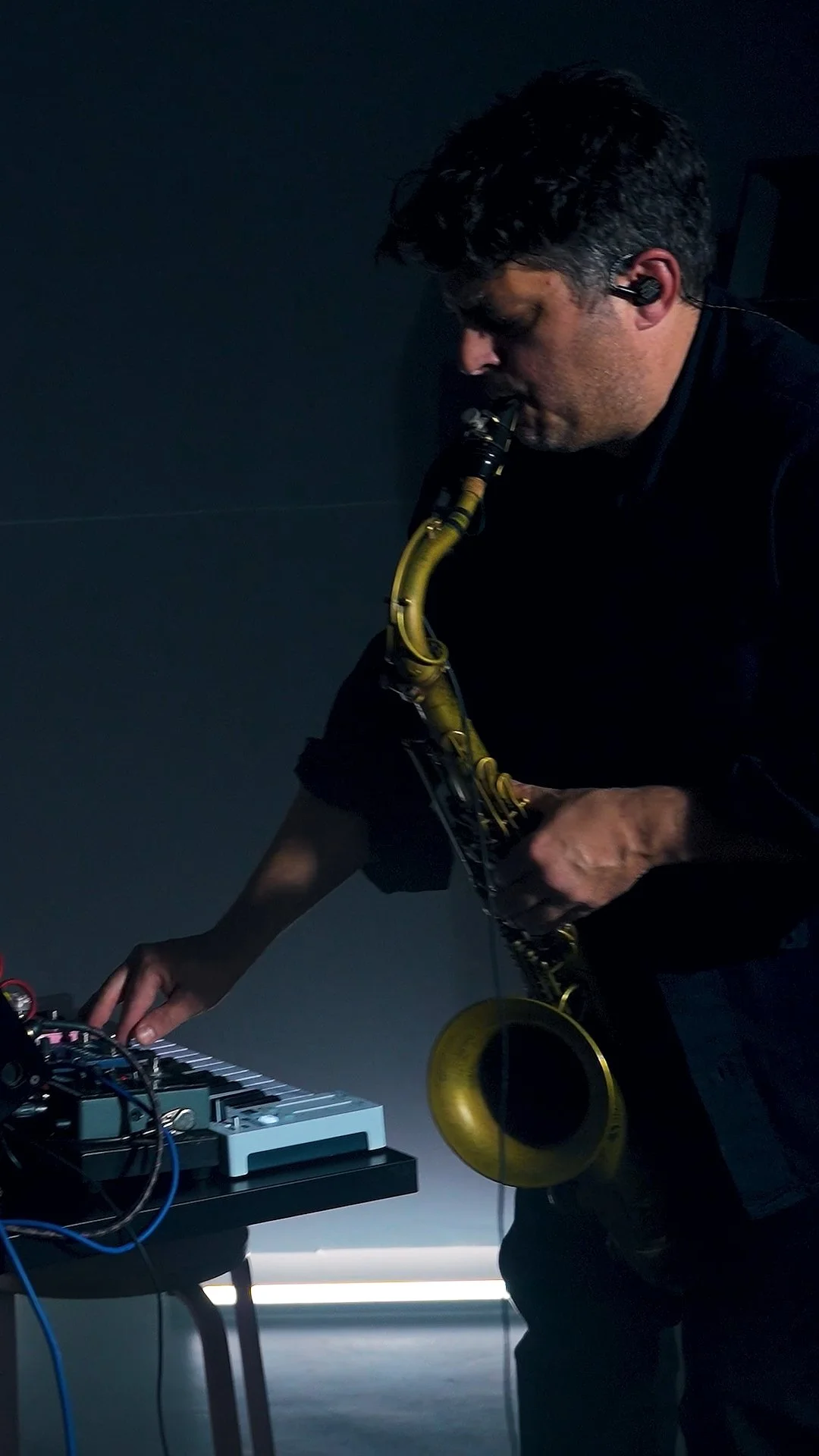Musician playing a yellow saxophone while operating electronic music equipment in a dimly lit setting.