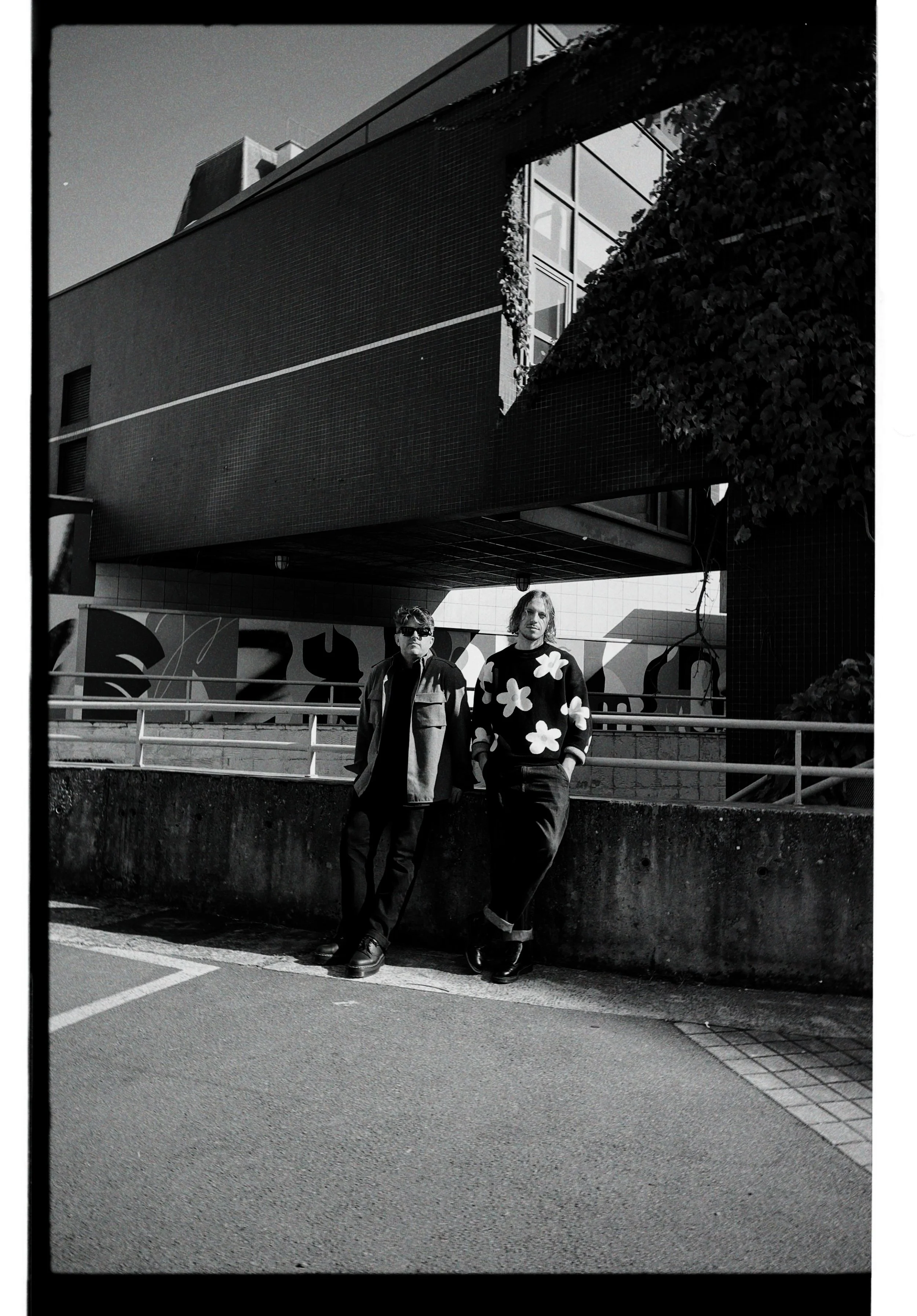 Two young men standing against a concrete barrier on a city street, with modern building and graffiti in the background, captured in black and white.