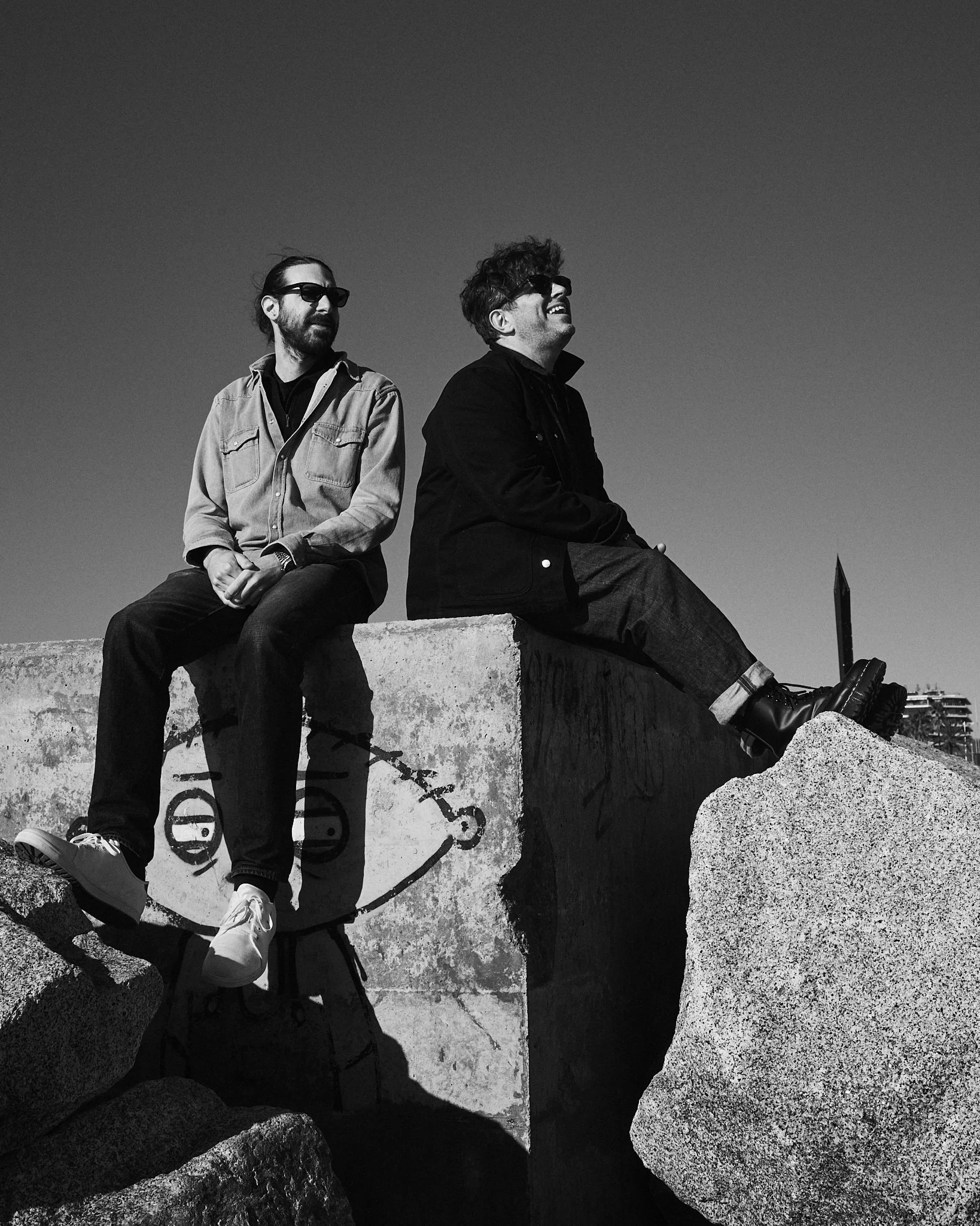 Two men wearing sunglasses sitting on concrete structures near the beach with rocks and a clear sky in the background.