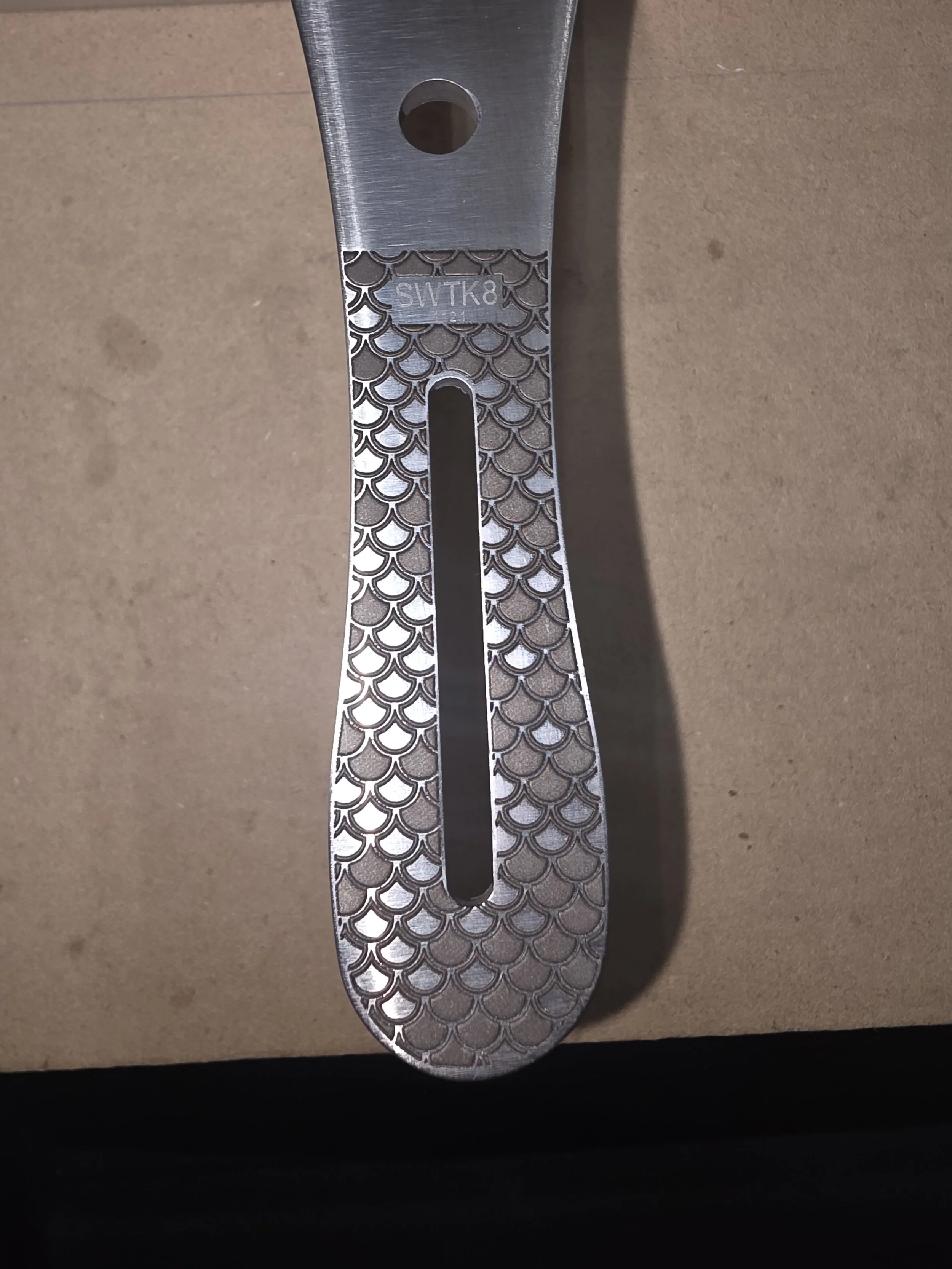 Metal folding ruler with a fish scale pattern design and a long central slot, placed on a brown surface.