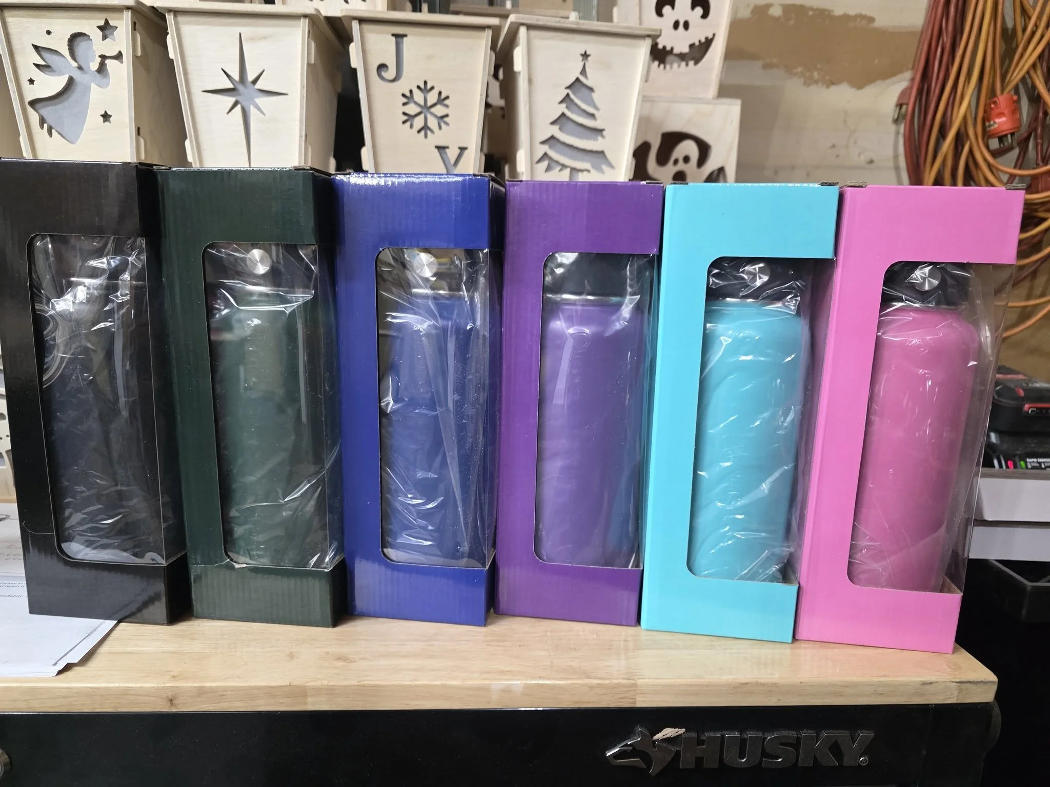 Six colorful candles in black, green, blue, purple, teal, and pink boxes on a wooden surface.
