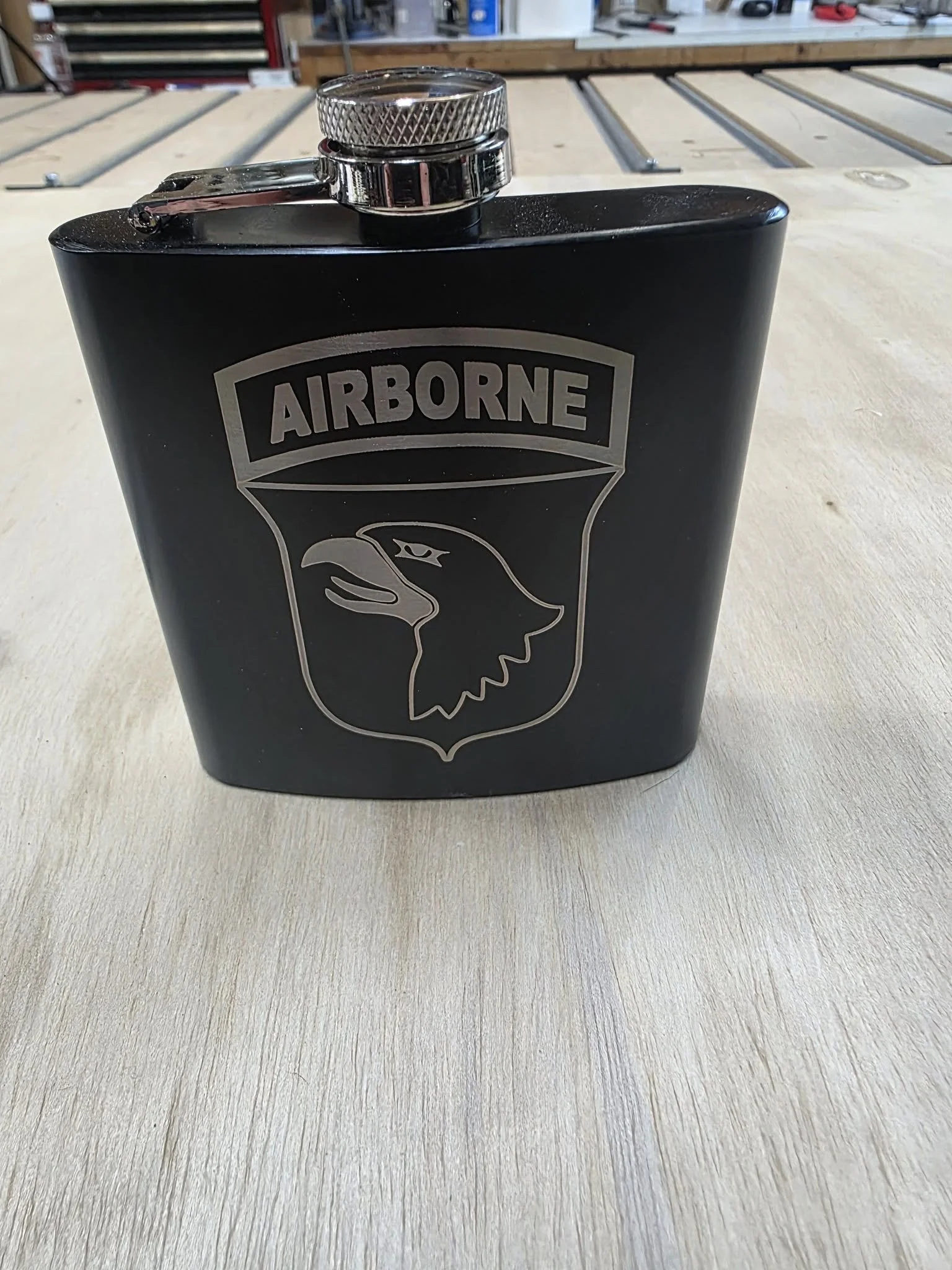 Black flask with an 'Airborne' military emblem featuring a duck head on a light wood surface in a workshop.