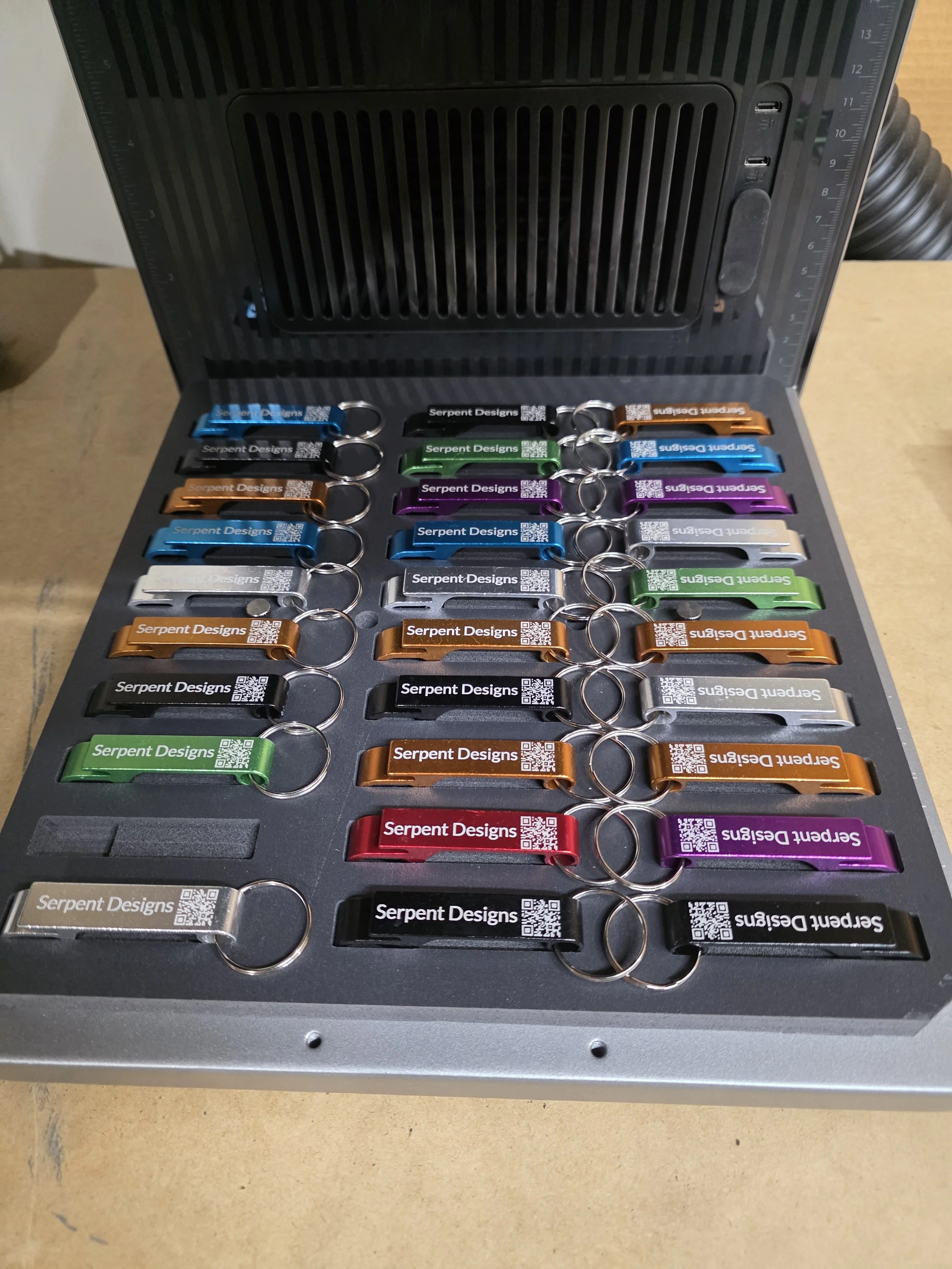 A collection of colorful keychains with 'Serpent Designs' printed on them, arranged inside a metal case with a black latch and a vent on top.