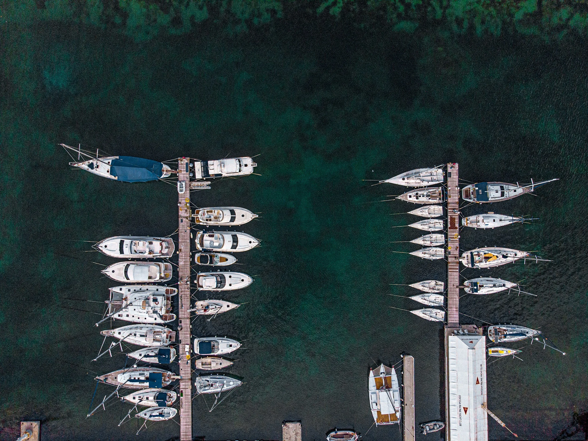 An aerial view of a marina with multiple boats docked along piers extending into the water. The boats vary in size and type, with some having covered decks and others open. The water appears dark green.