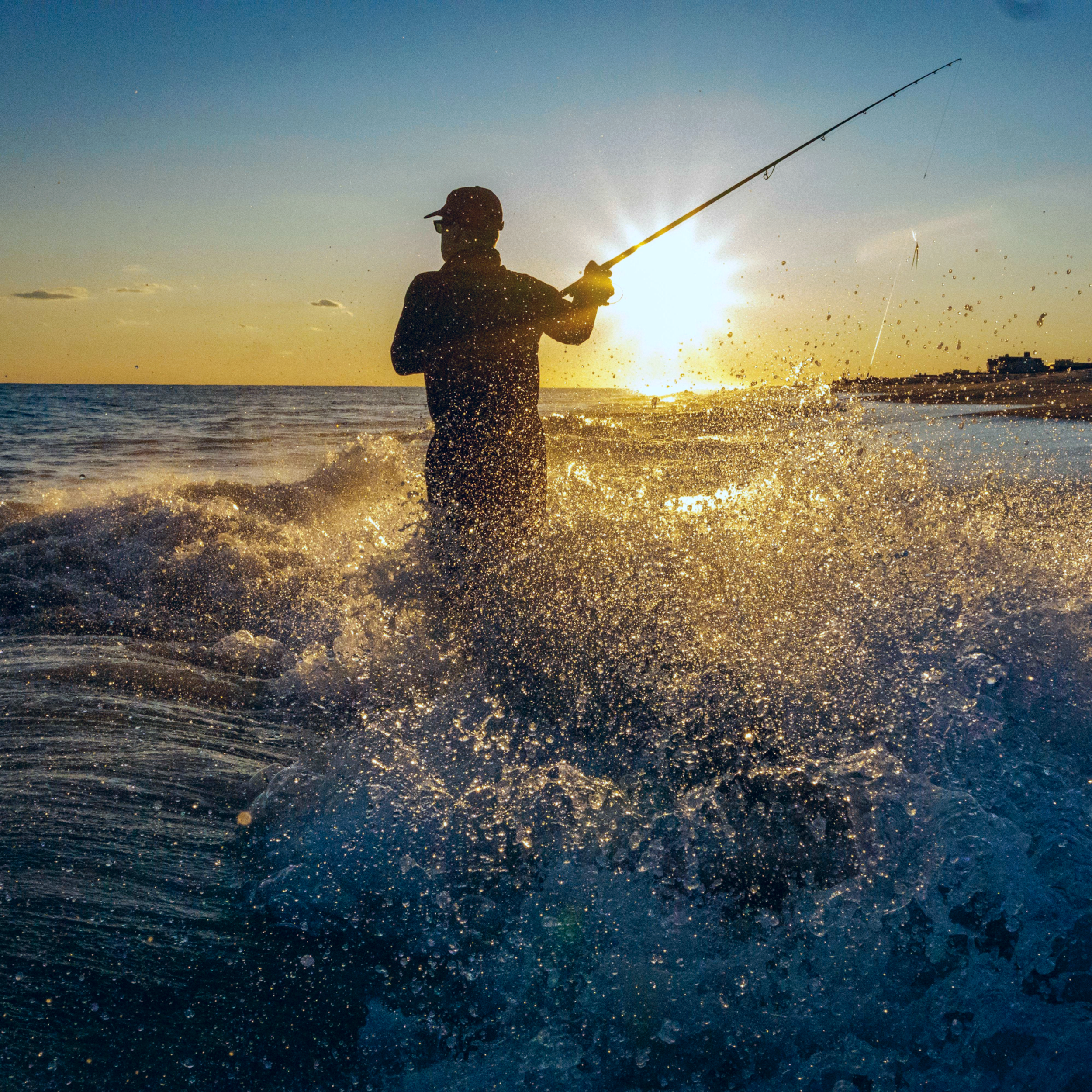 A person wearing a cap and sunglasses fishing on the shoreline with the sun setting in the background.