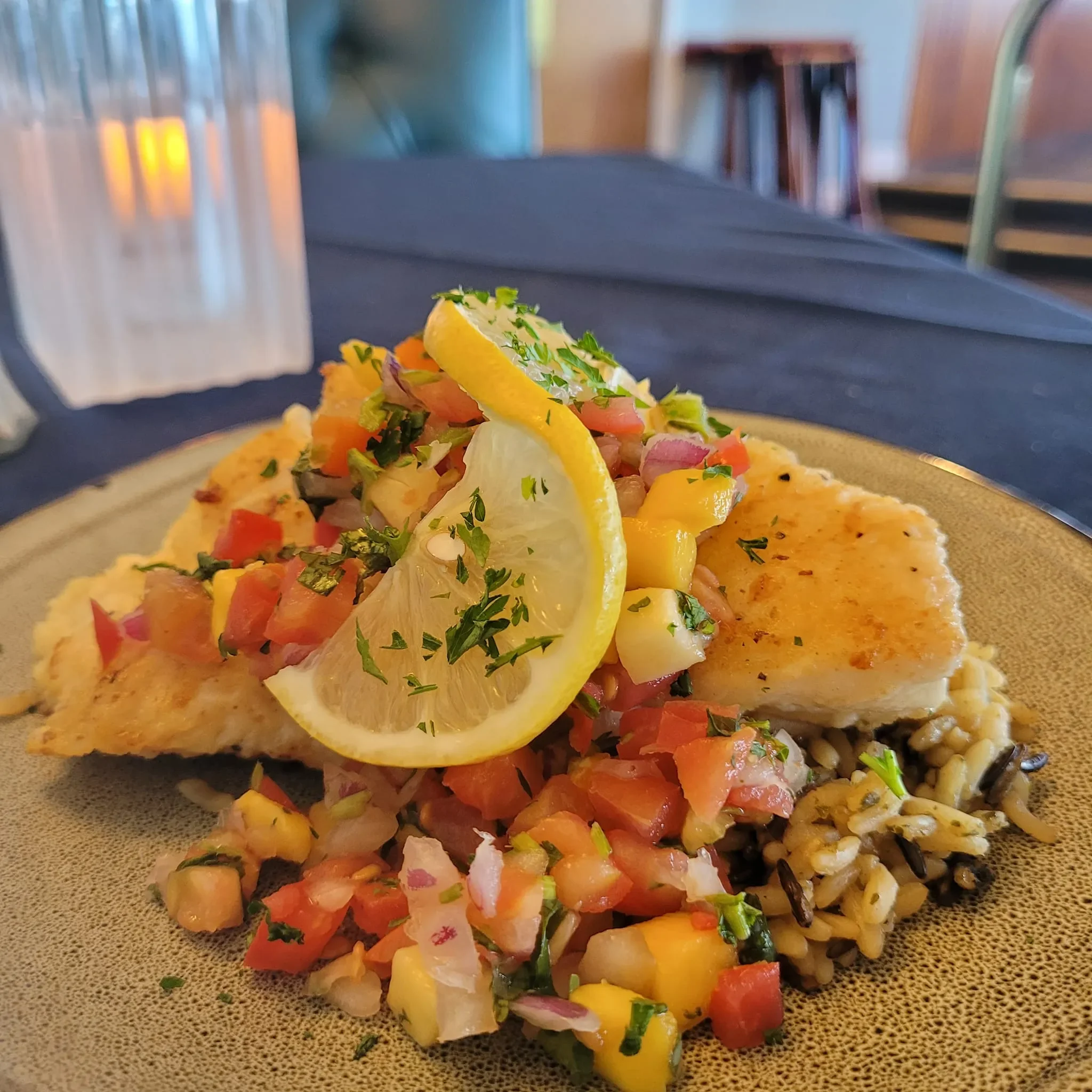 A plate with a piece of cooked white fish topped with lemon slices and chopped herbs, served with a salad of diced tomatoes, chopped onions, and mango, alongside a bed of mixed rice and beans.