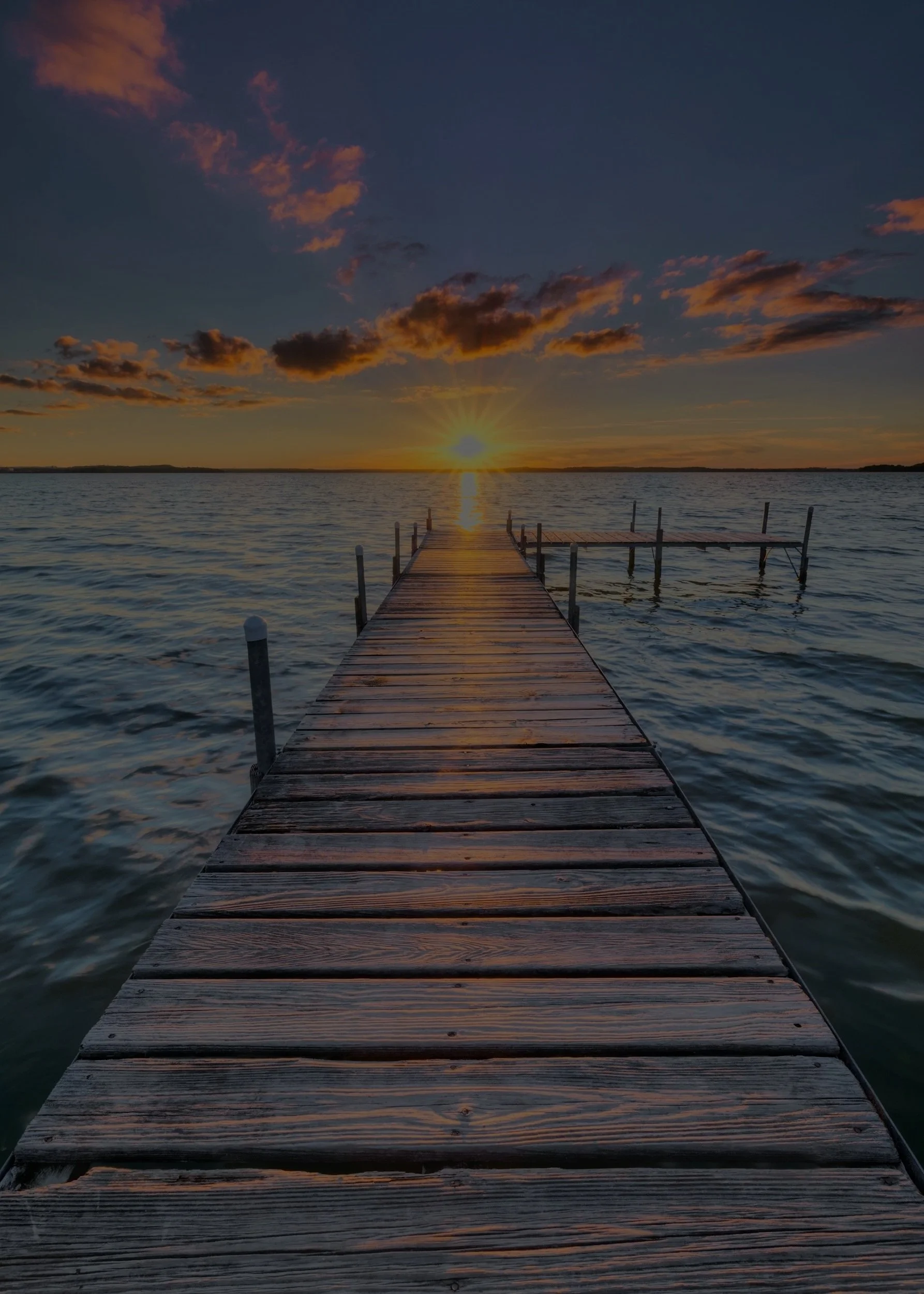 Wooden dock extending into a lake during sunset, with a few clouds in the sky and calm water surrounding the dock.