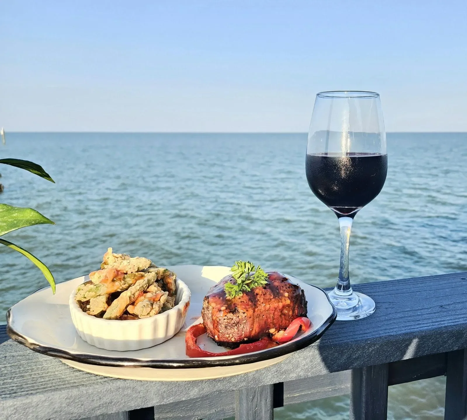 A plate with a baked meatloaf garnished with parsley and red peppers, a small bowl of fried vegetables, a glass of red wine, with a seaside view