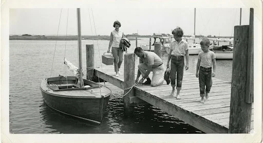 Four young boys on a wooden dock by a boat and water, with a woman standing further back on the dock, in a black and white photo.